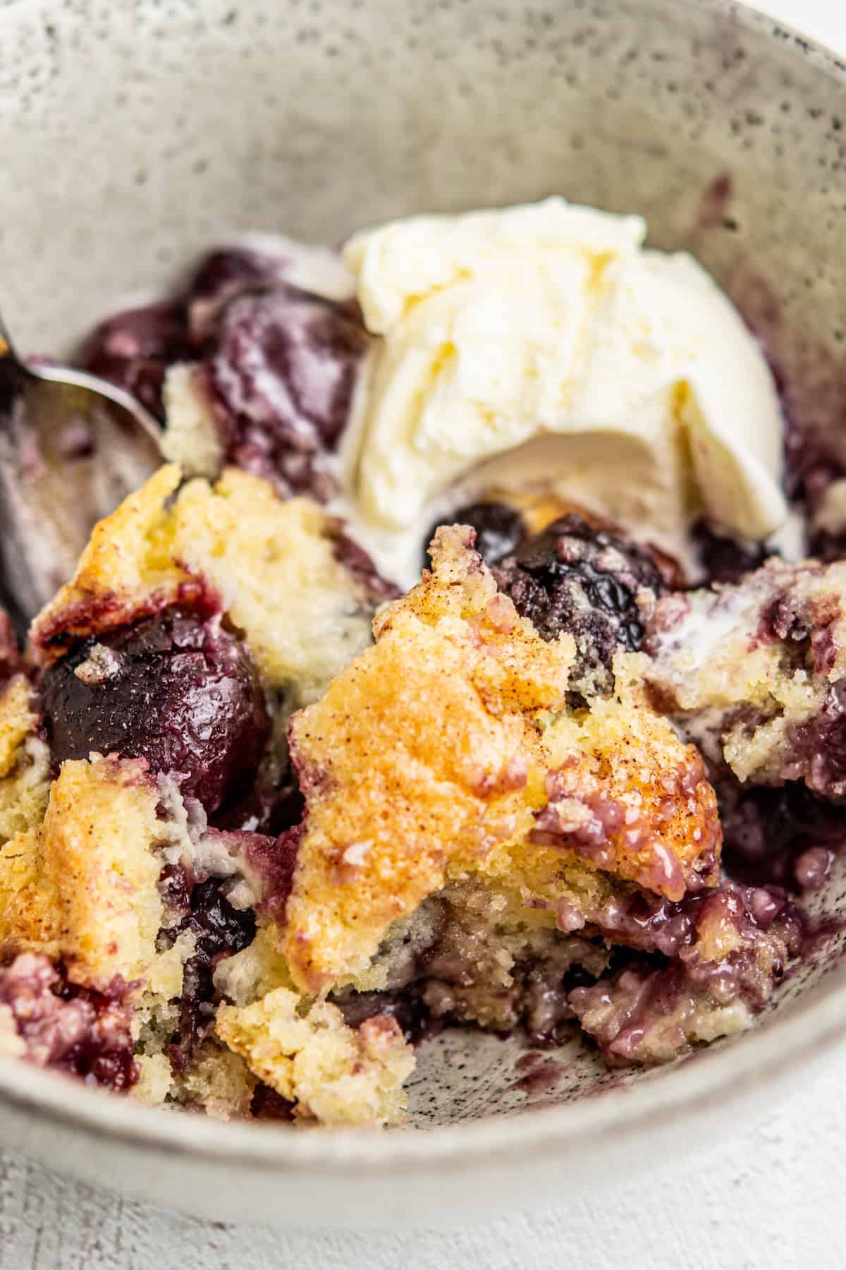 A close-up of a bowl containing a serving of cherry cobbler with a golden, crumbly topping, juicy cherries, and a scoop of vanilla ice cream melting on top. A spoon is partially visible on the side.