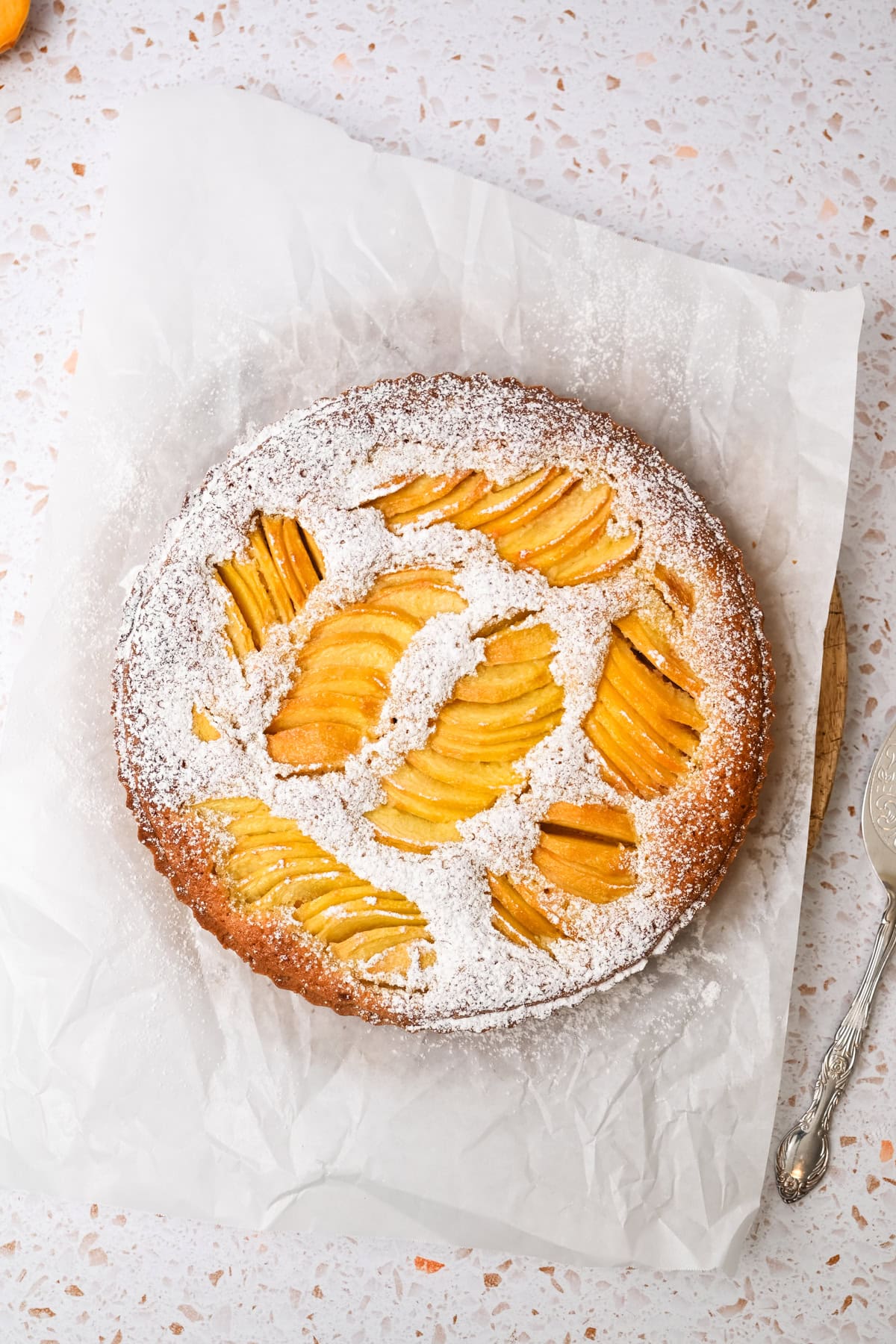 A round cake topped with sliced peaches and dusted with powdered sugar sits on white parchment paper. A vintage silver cake server rests nearby on a light, speckled surface.