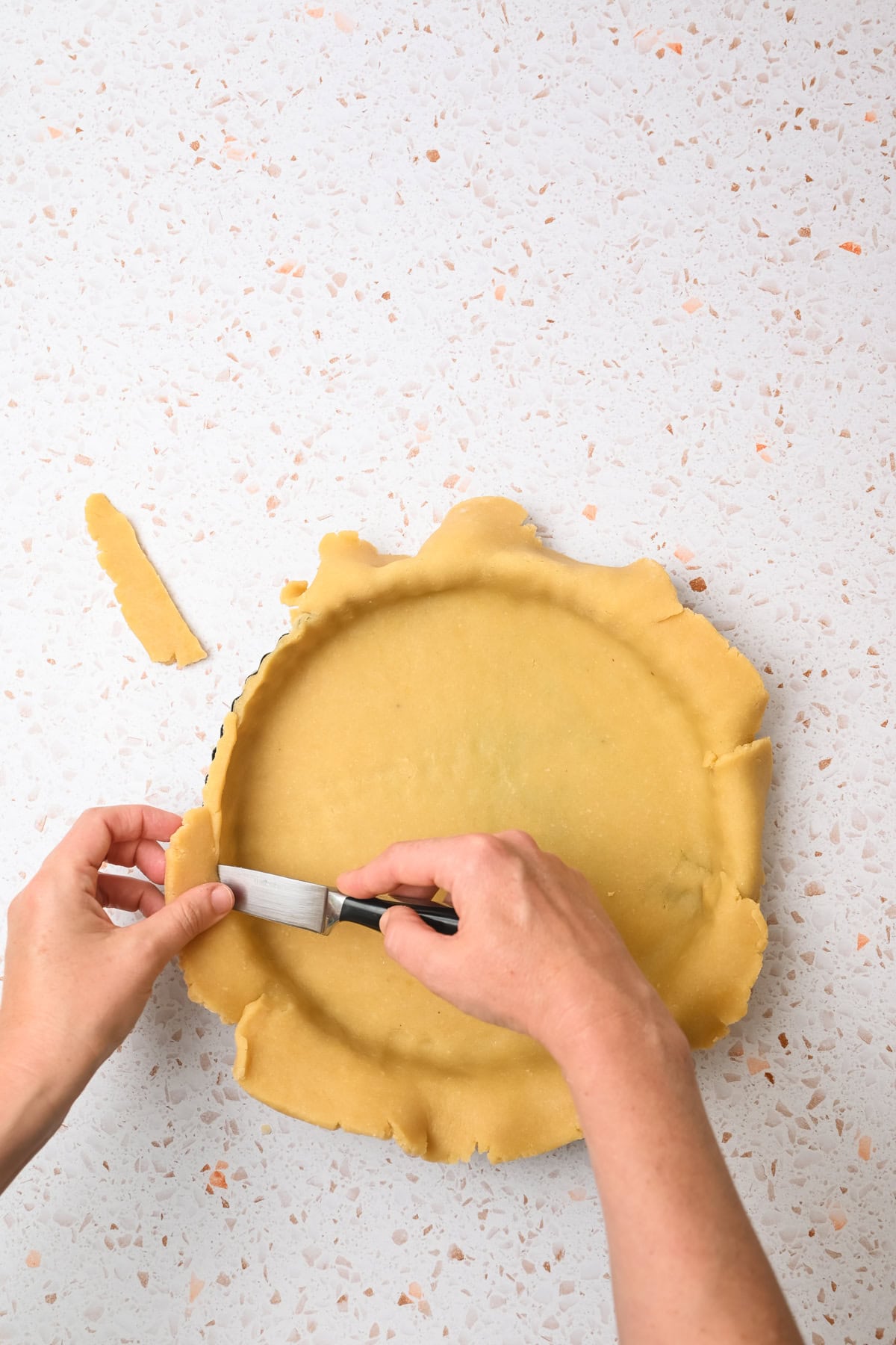 Two hands trim excess pie dough from the edge of a pie dish using a small knife on a light, speckled countertop. The uncooked dough is draped over the pie dish with uneven edges.