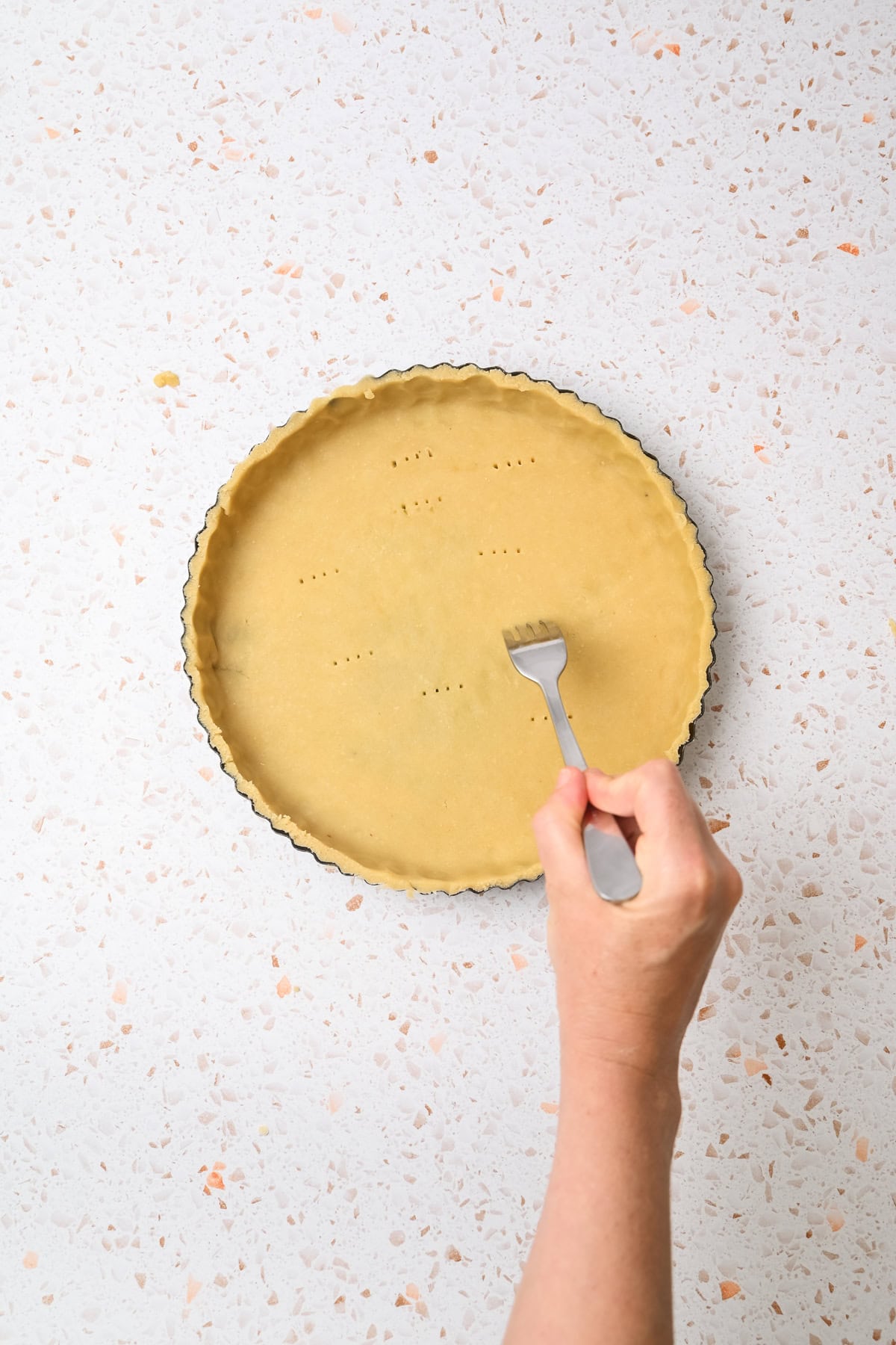 A hand holds a fork, poking holes in an unbaked tart crust in a round metal pan on a light speckled countertop.