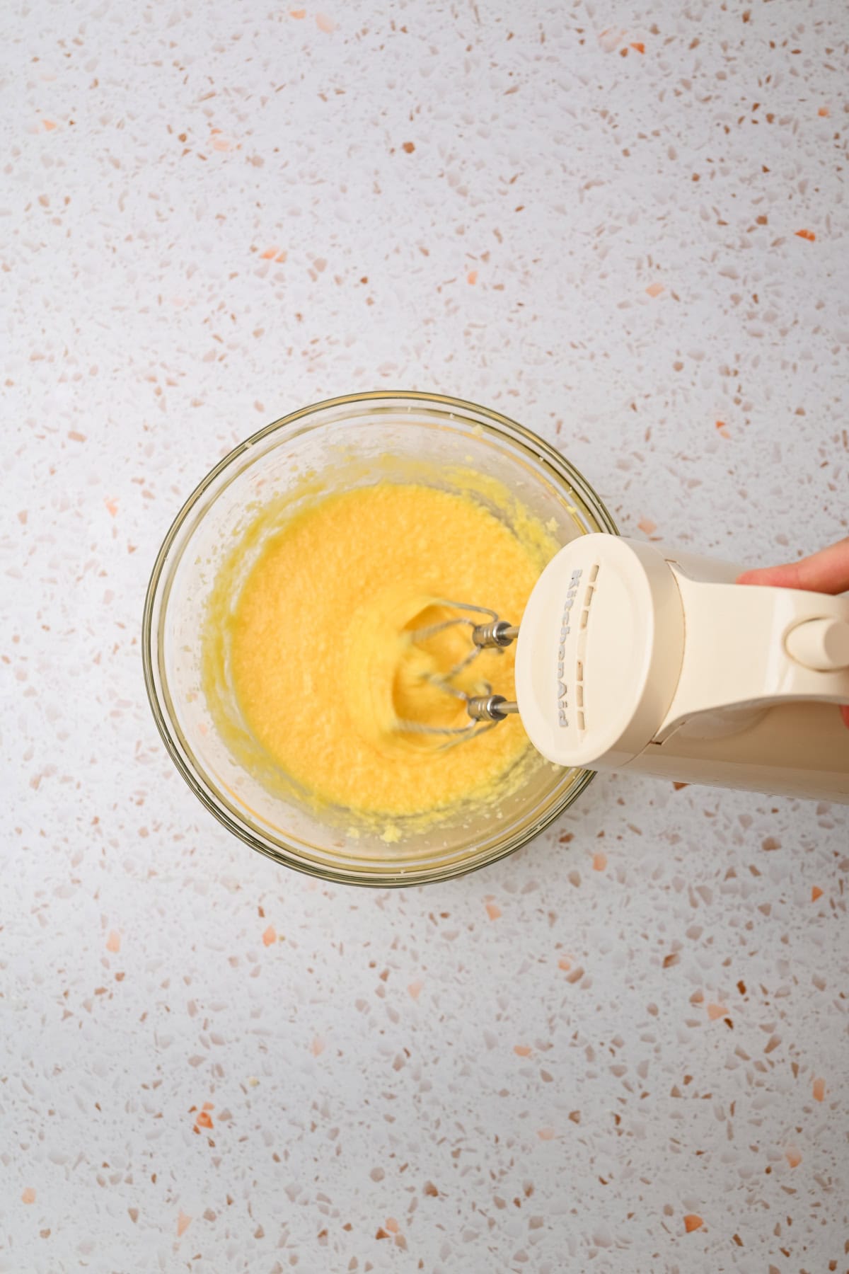 A hand holds an electric mixer blending a yellow mixture in a glass bowl on a speckled countertop, viewed from above.