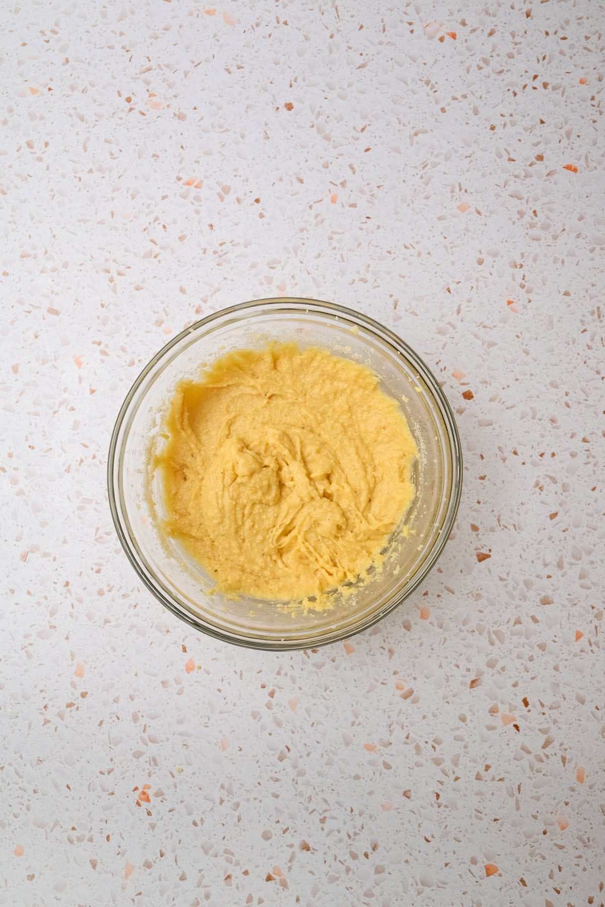 A glass bowl filled with creamy, light yellow batter sits on a white speckled countertop.