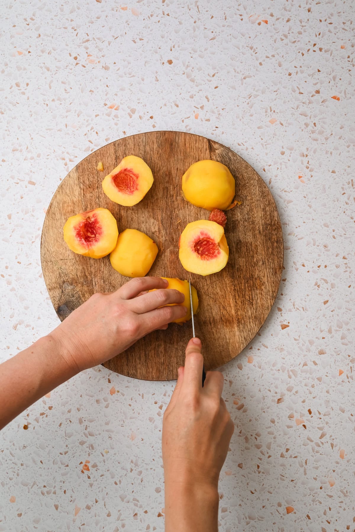 Two hands slice a peeled peach on a round wooden cutting board with more peeled and halved peaches on it, placed on a speckled countertop.