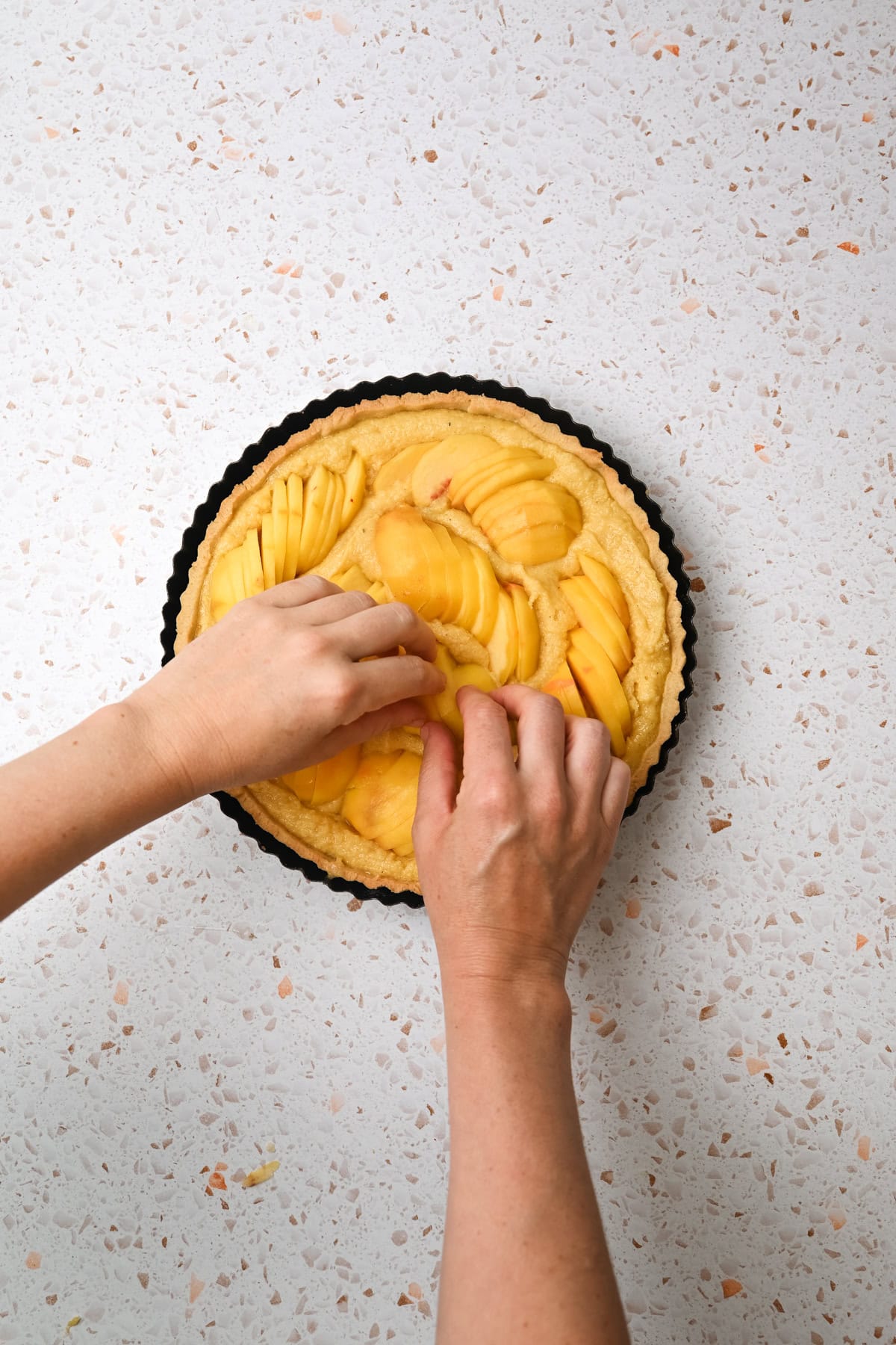 A pair of hands arranging thin slices of yellow fruit, possibly mango or peach, on top of a tart crust in a round, fluted pan, set on a light terrazzo countertop.