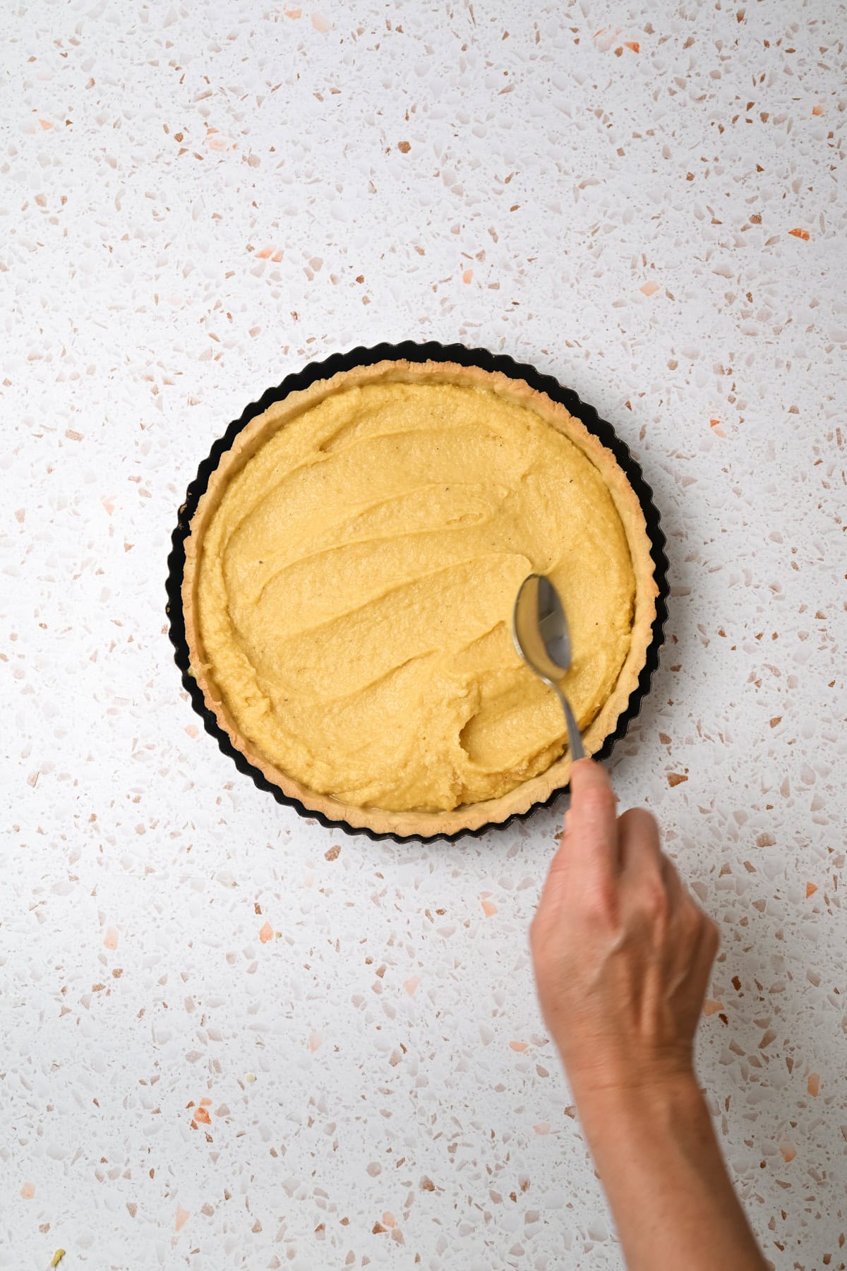 A hand holds a spoon, spreading a yellow, creamy filling evenly into a round tart crust on a light speckled countertop.