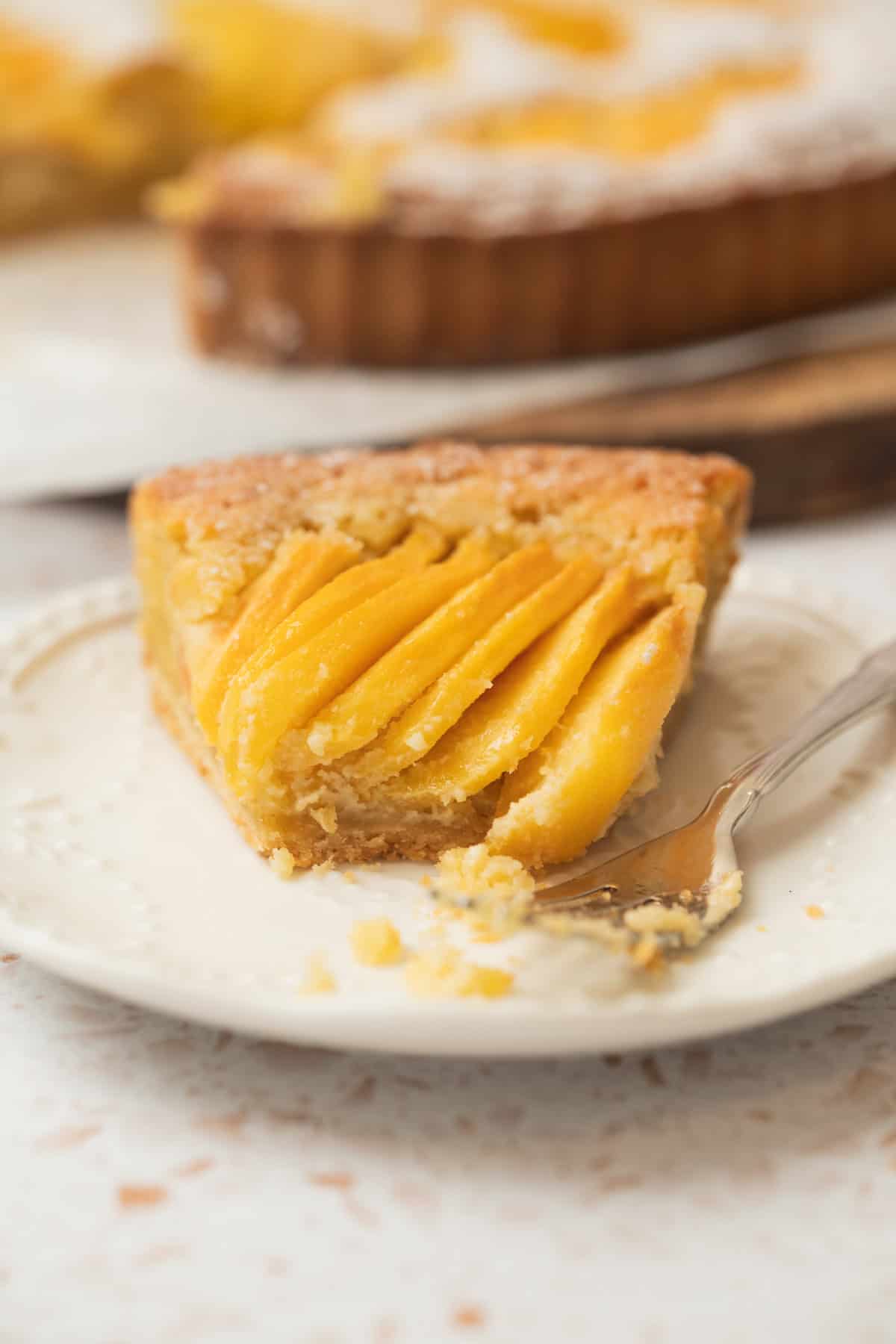 A close-up of a slice of mango tart on a white plate with a fork, showing layers of golden mango slices on top of a crumbly crust. The background shows the rest of the tart, slightly out of focus.