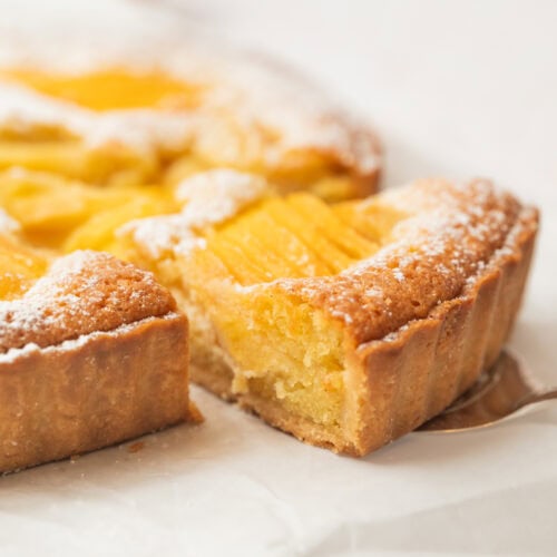 A close-up of a peach tart with a golden crust, topped with peach slices and dusted with powdered sugar. One slice has been cut and is slightly pulled away from the tart, showing its moist interior.