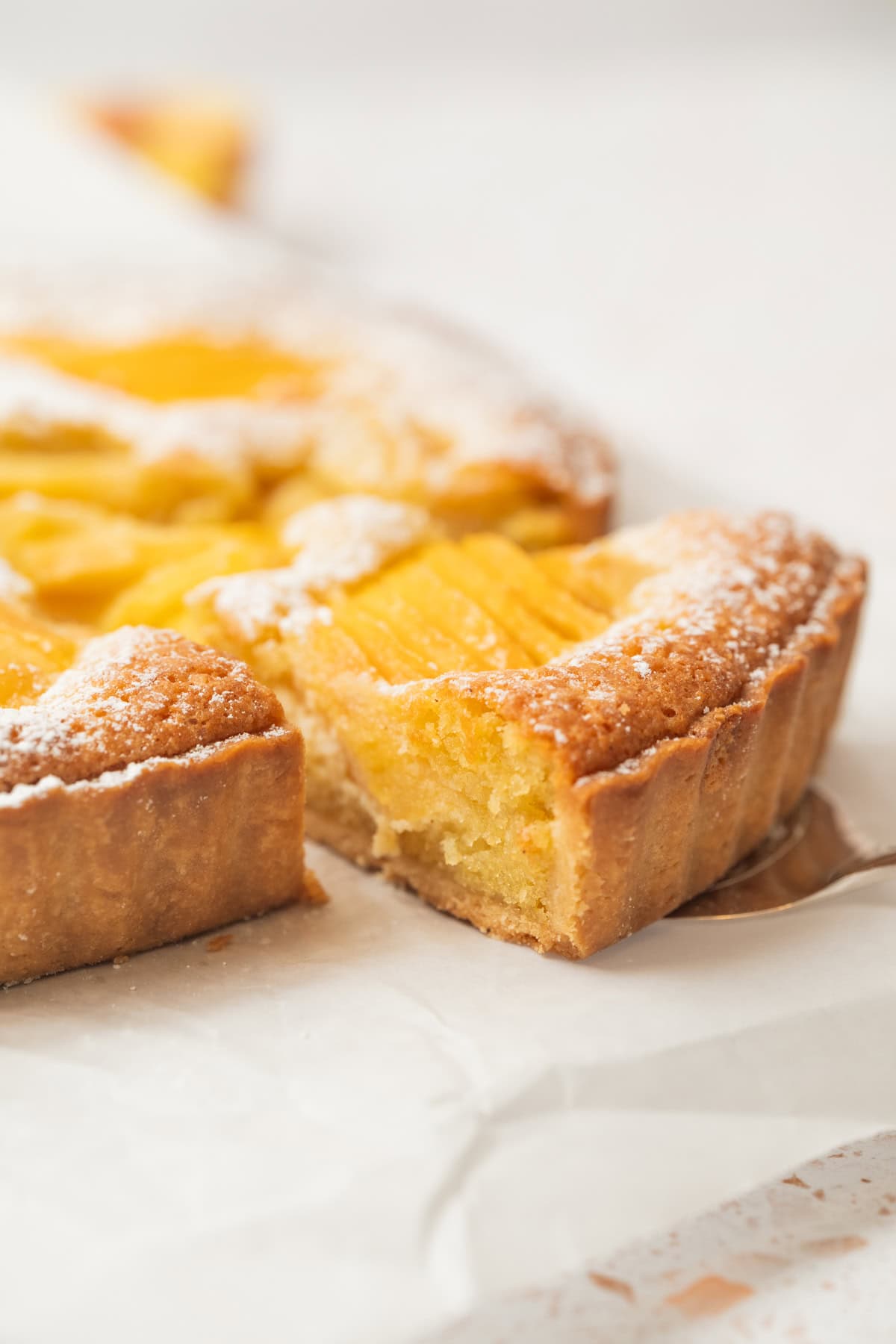 A close-up of a peach tart with a golden crust, topped with peach slices and dusted with powdered sugar. One slice has been cut and is slightly pulled away from the tart, showing its moist interior.