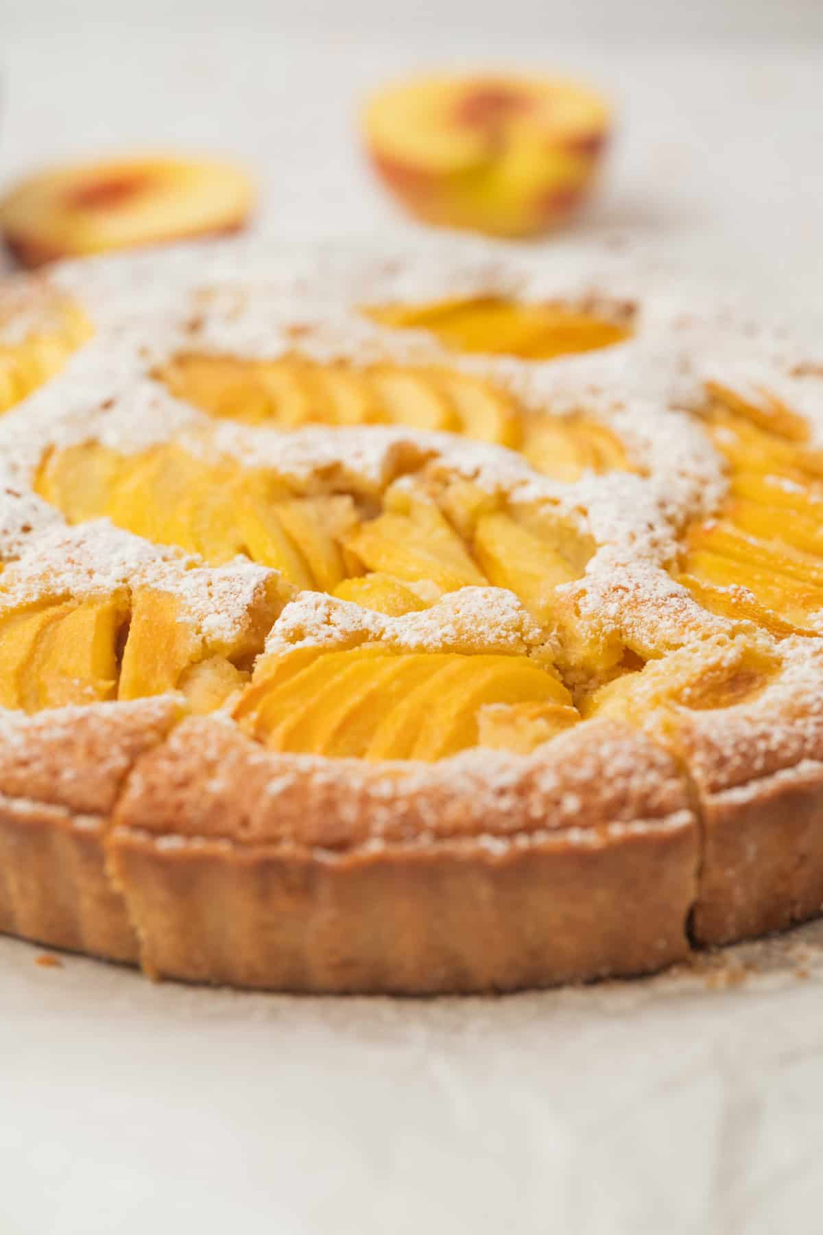 A close-up of a peach tart topped with sliced fresh peaches and dusted with powdered sugar, with blurred peach halves in the background.