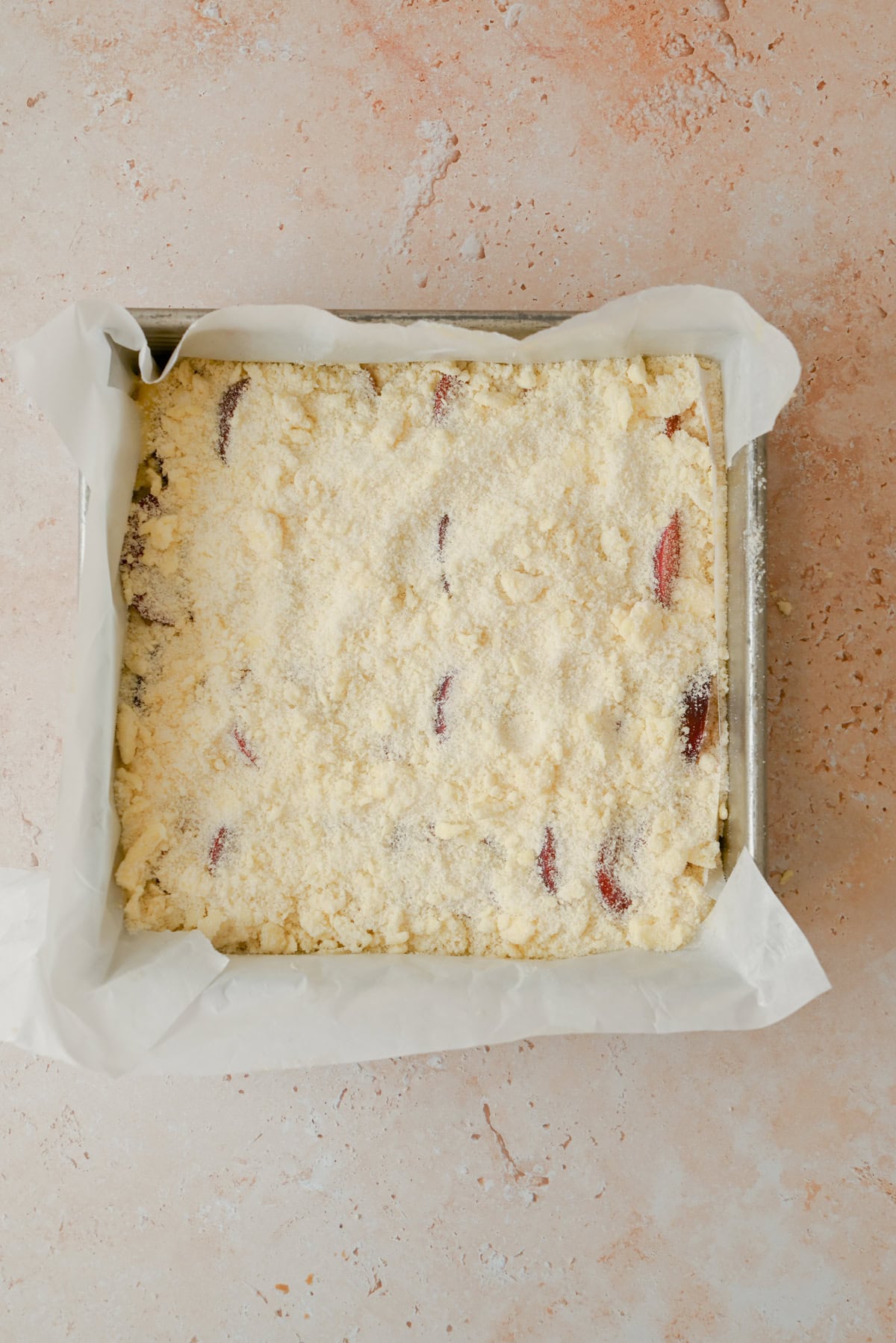 A square baking pan lined with parchment paper, filled with an unbaked crumbly dough mixture. Pieces of fruit, possibly plums, are partially visible beneath the crumb topping. The pan is on a lightly textured surface.