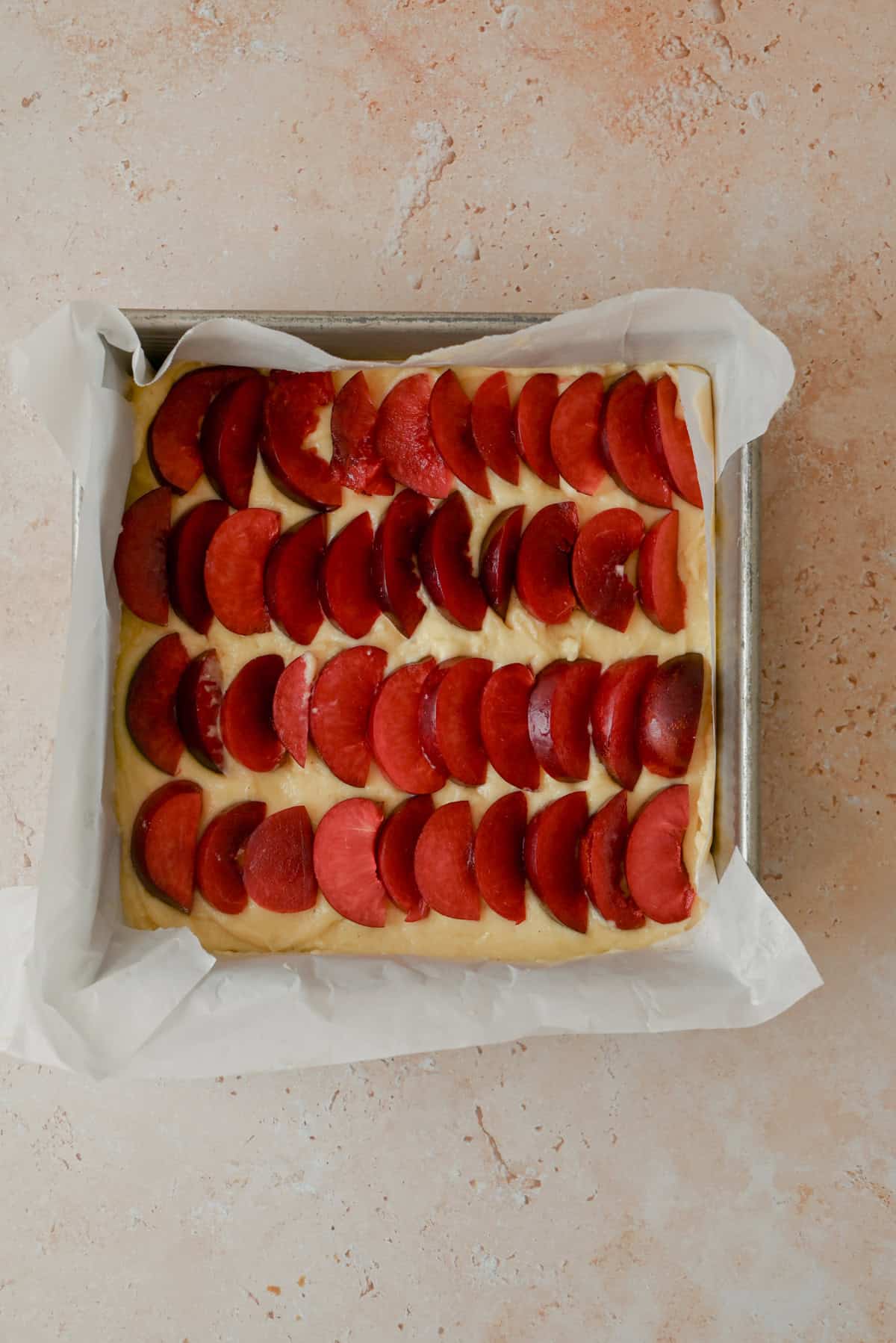 A rectangular baking pan lined with parchment paper holds a baked dessert topped with evenly arranged slices of red plums on a light, textured surface.