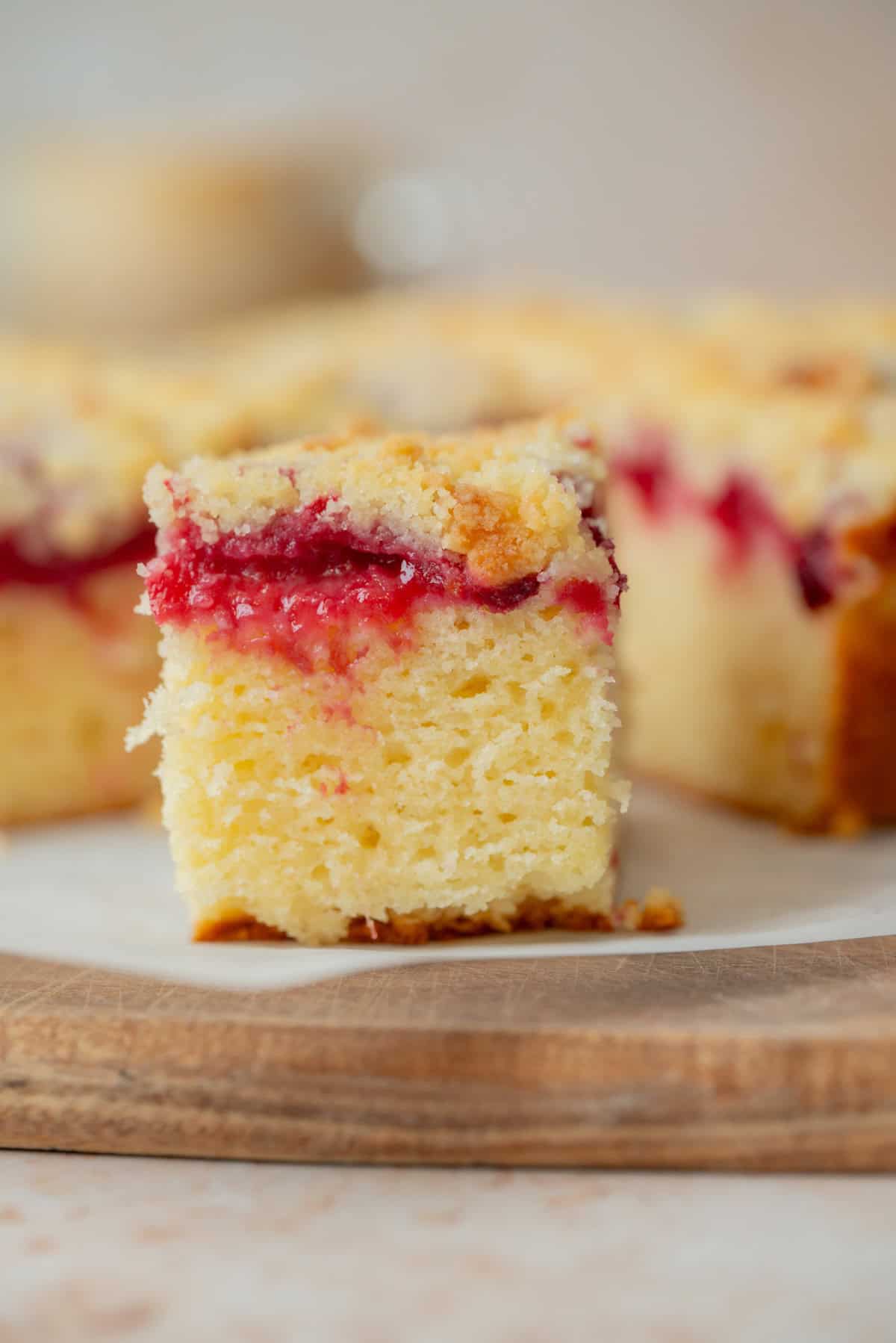 A close-up of a square slice of yellow plum cake with a layer of red fruit filling and crumb topping, set on a wooden board with more cake pieces blurred in the background.
