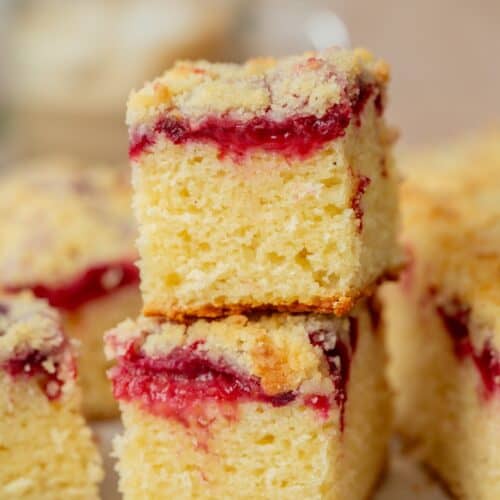 Two square slices of crumbly yellow plum yoghurt cake with a layer of red fruit jam in the middle, stacked on top of each other, with more pieces in the background.