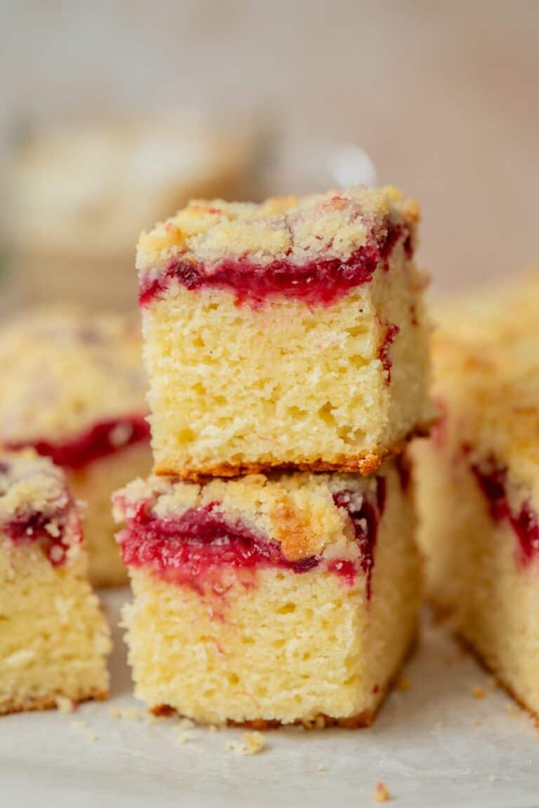 Two square slices of crumbly yellow plum yoghurt cake with a layer of red fruit jam in the middle, stacked on top of each other, with more pieces in the background.