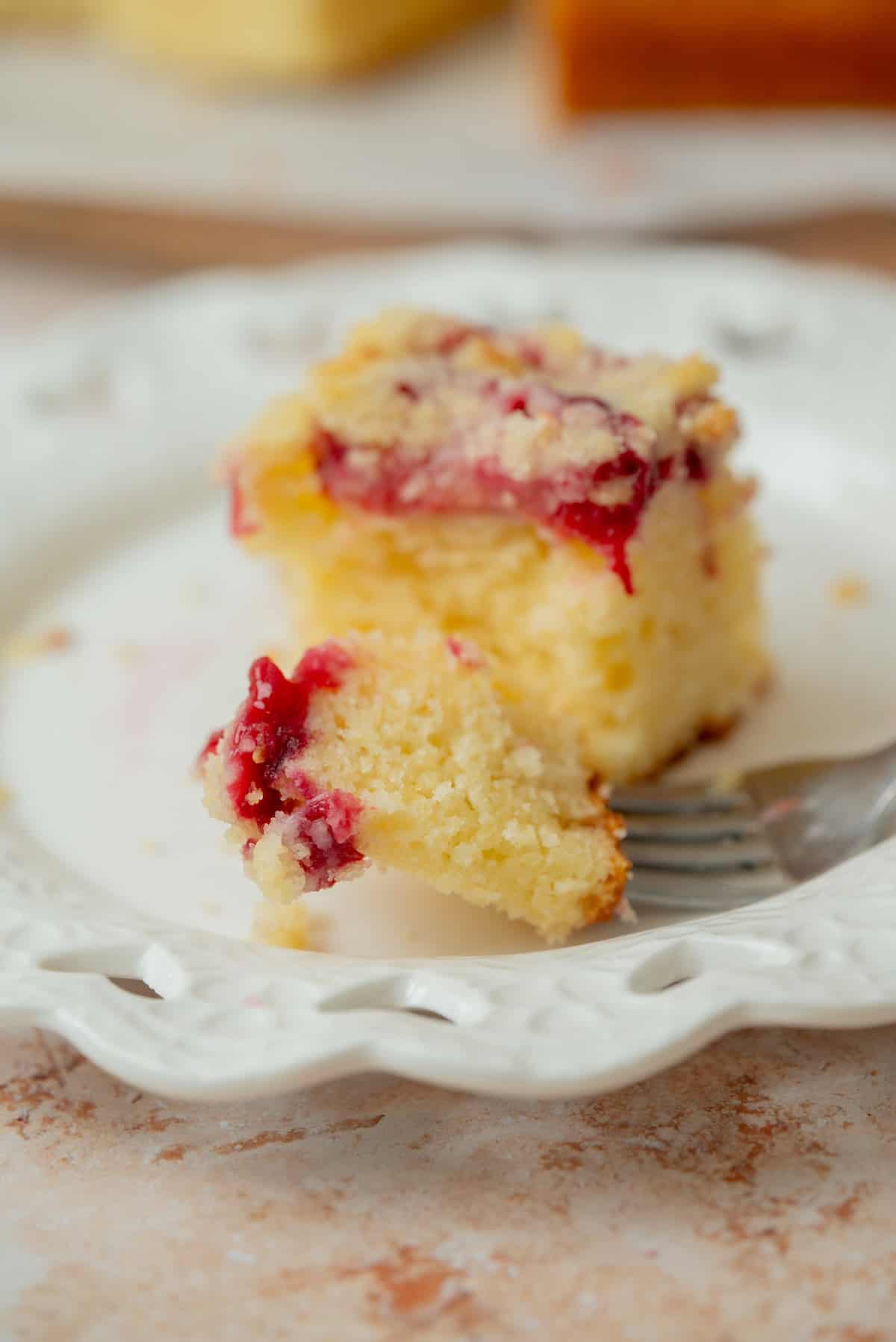 A close-up of a piece of crumb-topped plum yogurt cake with a layer of red fruit, partially eaten, on a decorative white plate with a fork resting nearby.