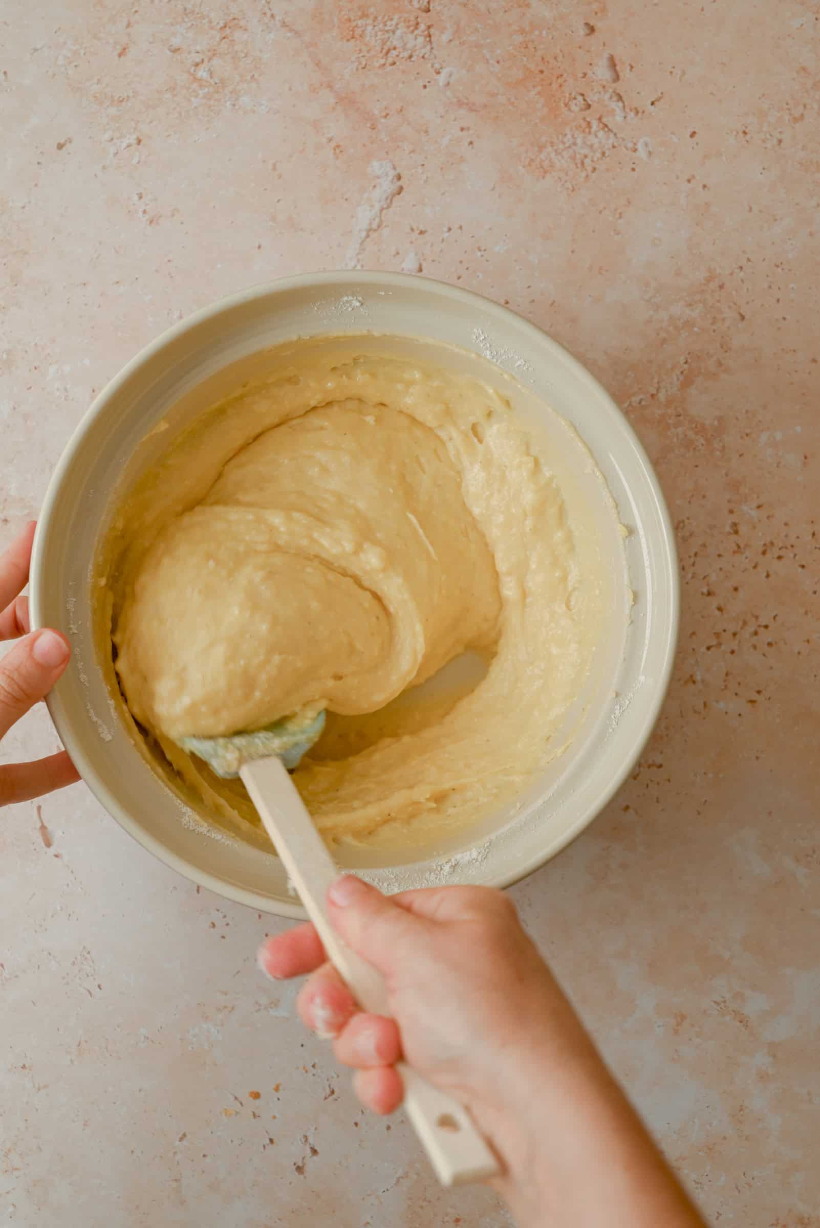A person mixes thick yellow cake batter in a beige bowl using a spatula, set on a light speckled countertop.