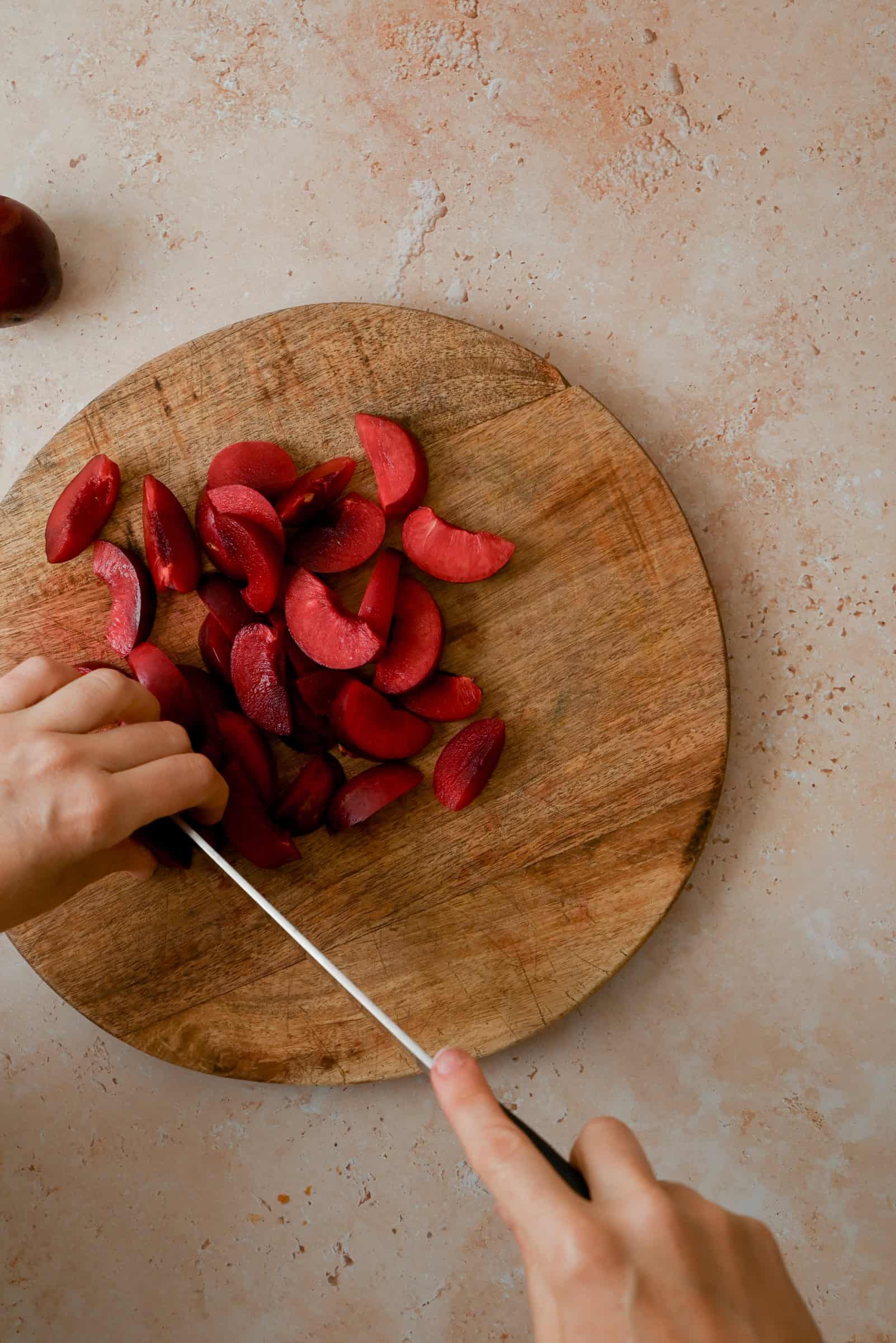 A person slices red plums on a round wooden cutting board, with several plum wedges already cut, on a light-colored countertop.