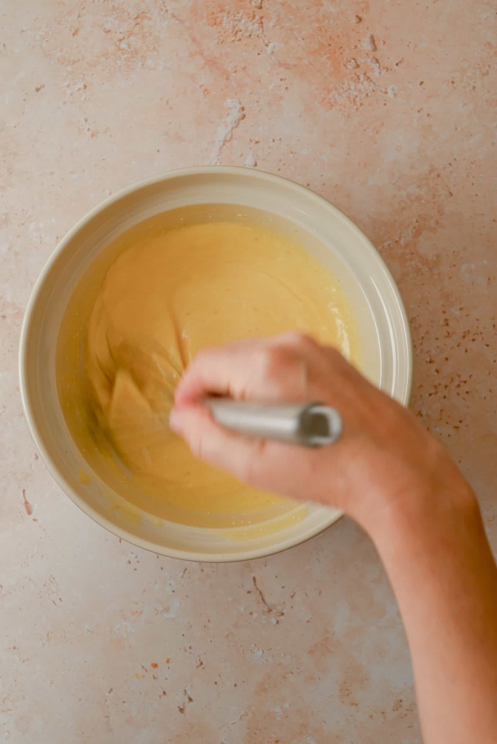 A hand is whisking a creamy yellow batter in a beige bowl on a light, speckled countertop.