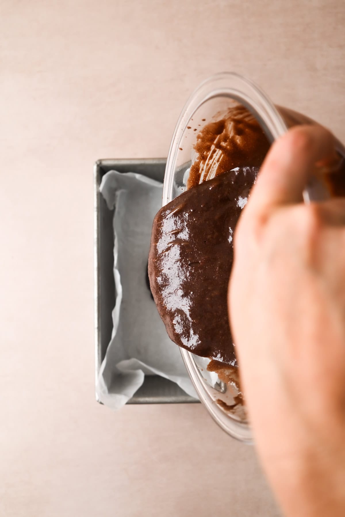A hand pours chocolate cake batter from a glass bowl into a parchment-lined loaf pan on a light-colored surface.