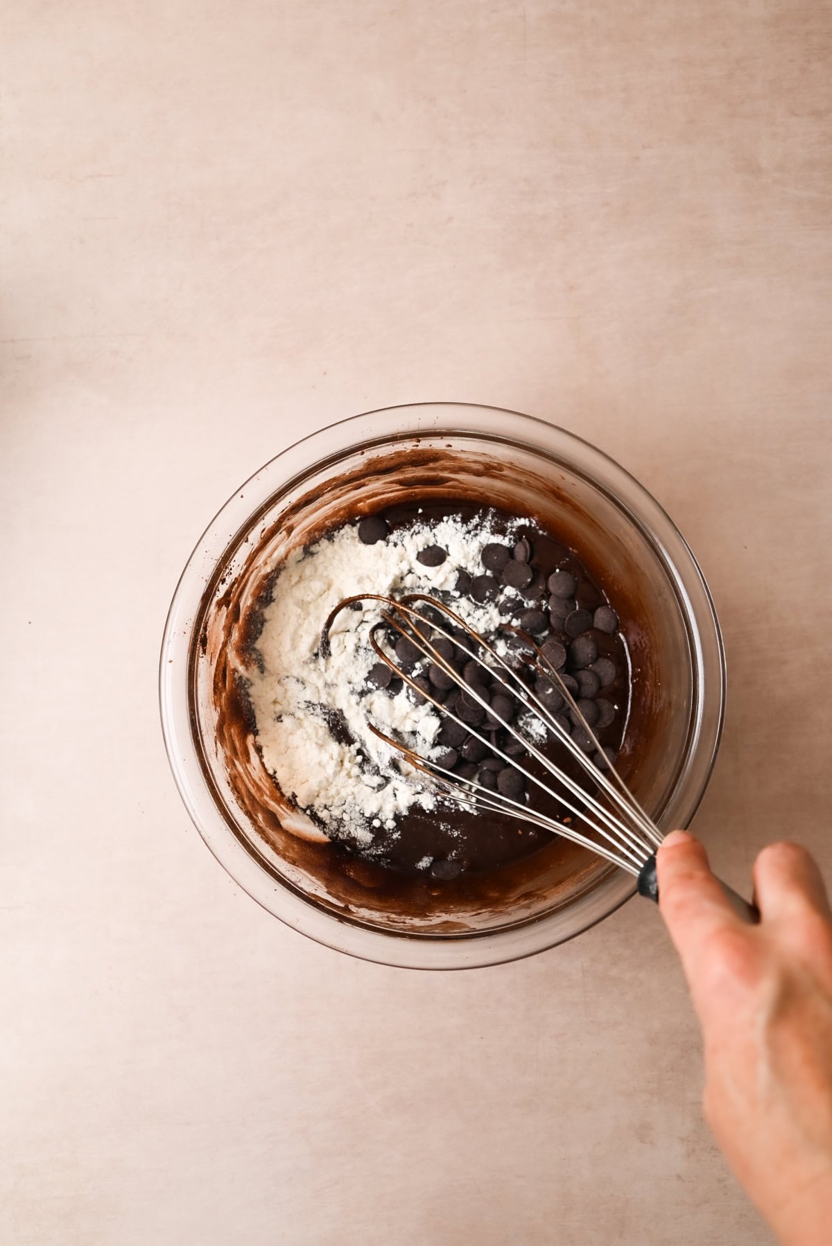 A hand holding a whisk mixes flour and chocolate chips into a bowl of chocolate batter on a light-colored surface.