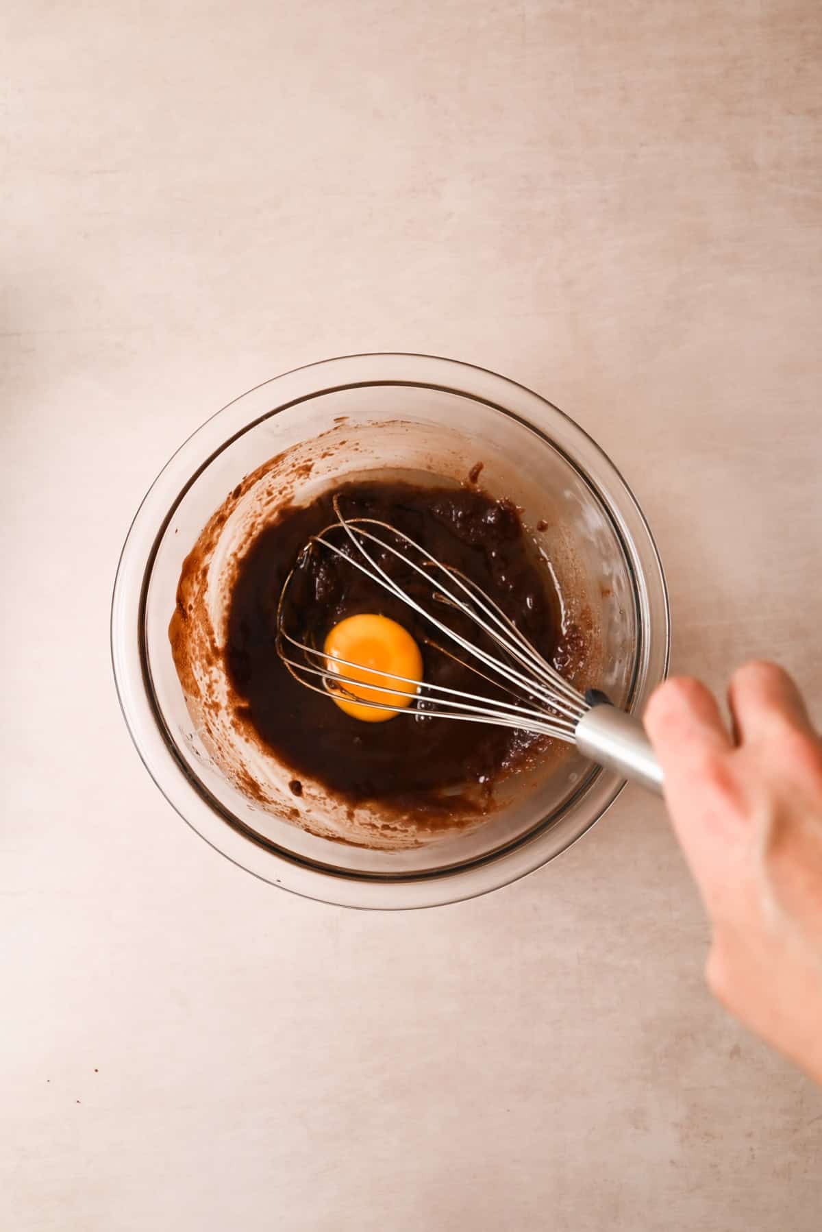 A hand holds a whisk above a glass bowl containing brownie batter and a raw egg, ready to be mixed, on a light countertop.