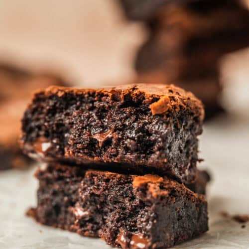 Two rich, fudgy chocolate brownies are stacked on a piece of parchment paper, showing a moist, gooey interior and a slightly crisp top. More brownies are blurred in the background.