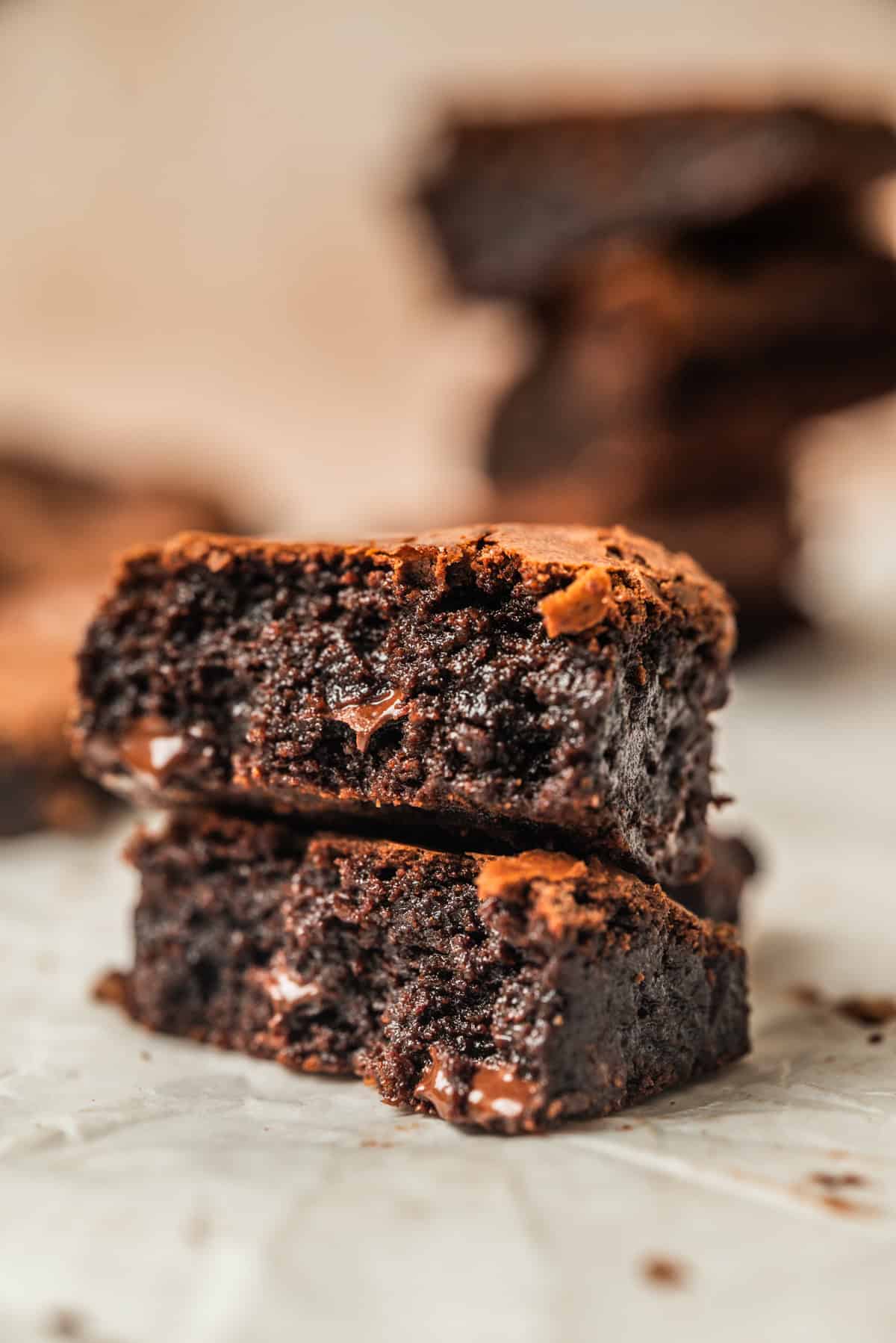 Two rich, fudgy chocolate brownies are stacked on a piece of parchment paper, showing a moist, gooey interior and a slightly crisp top. More brownies are blurred in the background.