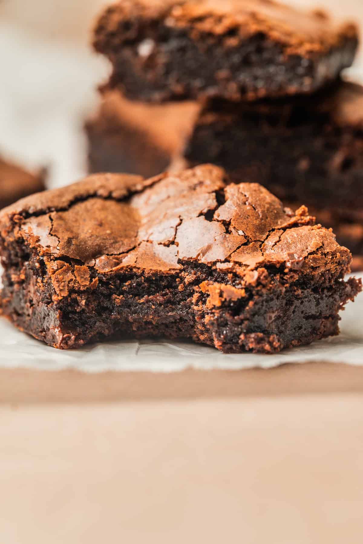 A close-up of a fudgy chocolate brownie with a cracked top and a bite taken out, resting on parchment paper with more brownies blurred in the background.