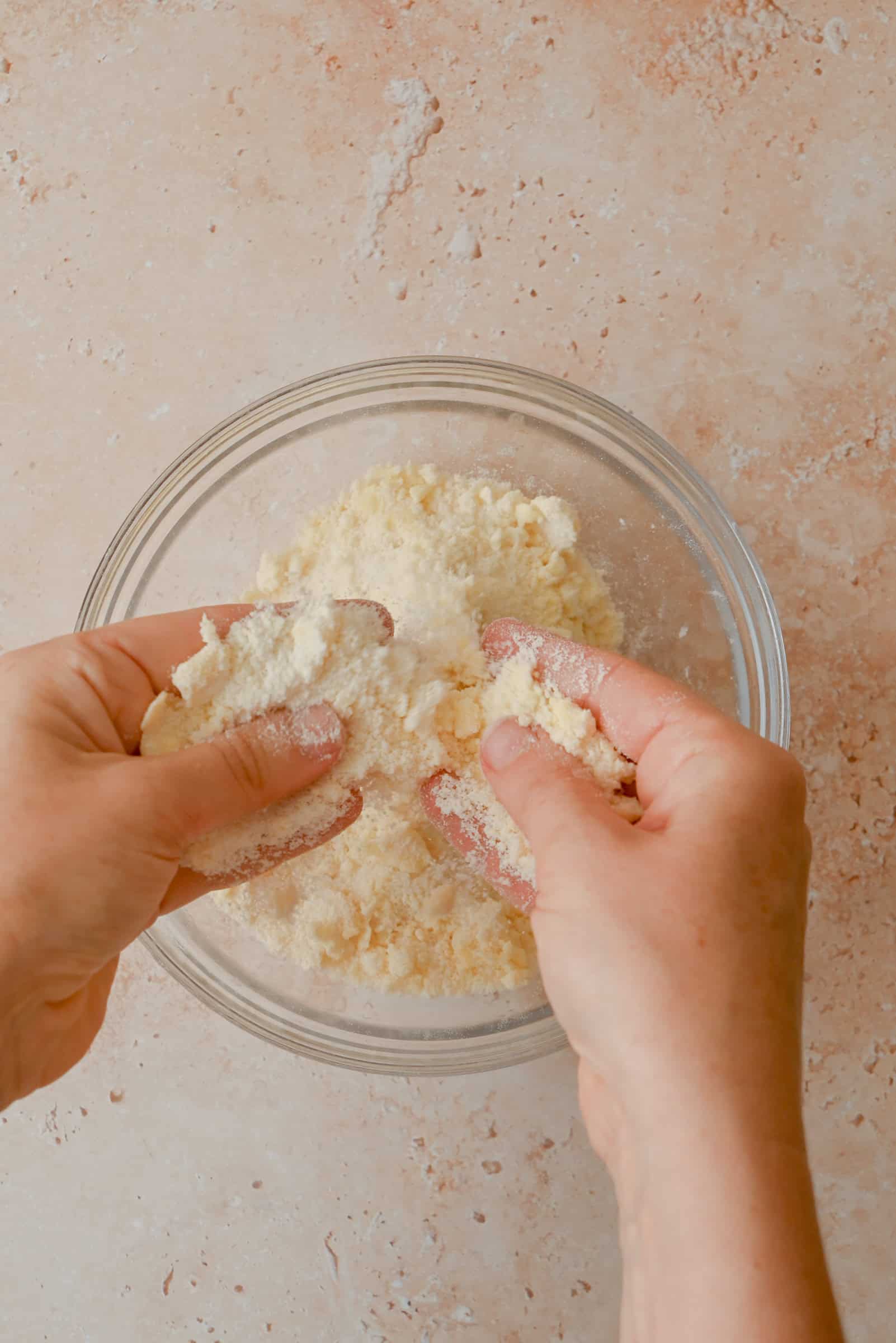 Two hands crumble a mixture of flour and butter in a glass bowl on a beige countertop, preparing a dough or pastry base.