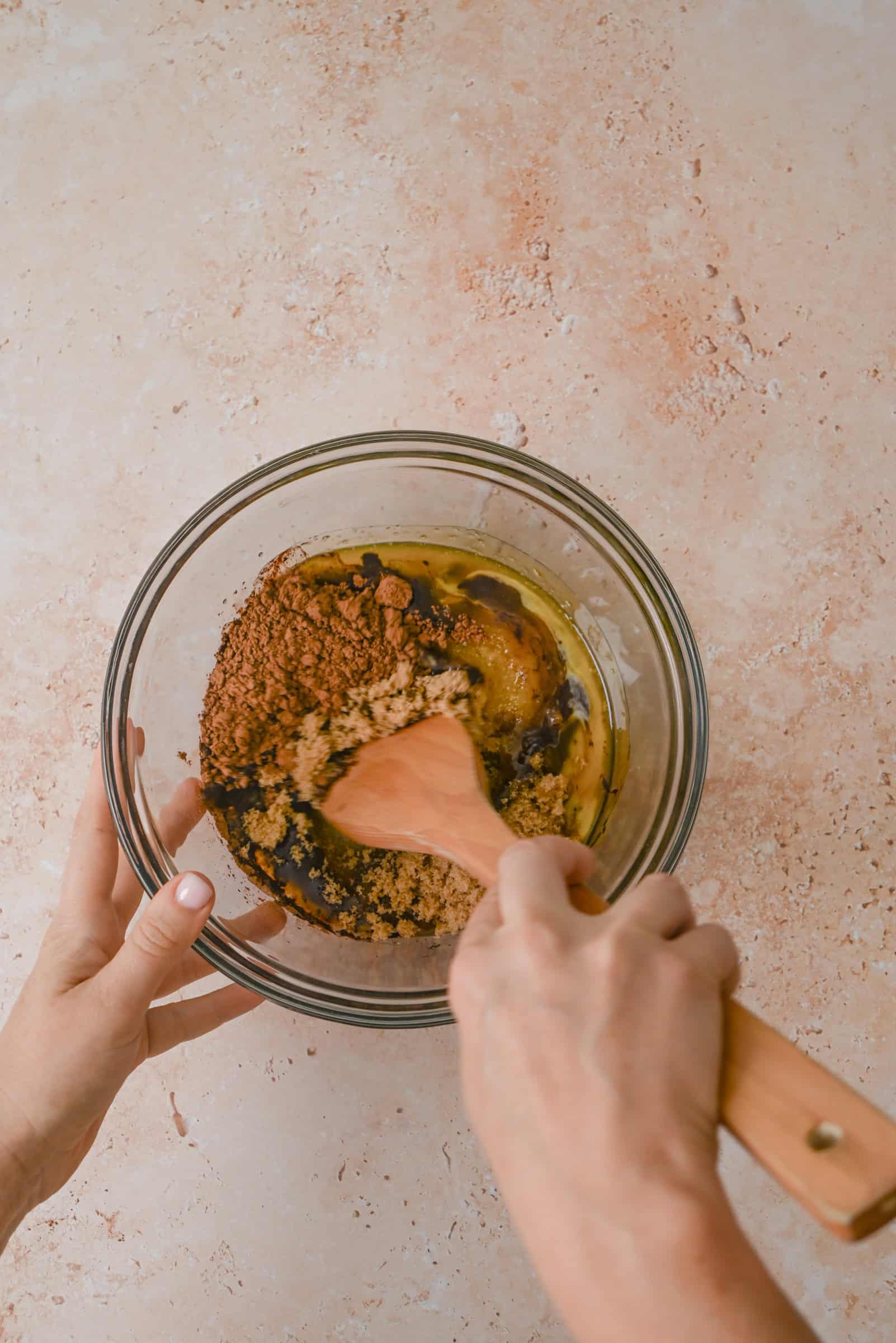 Hands stirring brown sugar, cocoa powder, eggs, and oil in a glass mixing bowl with a wooden spoon on a light-colored countertop.