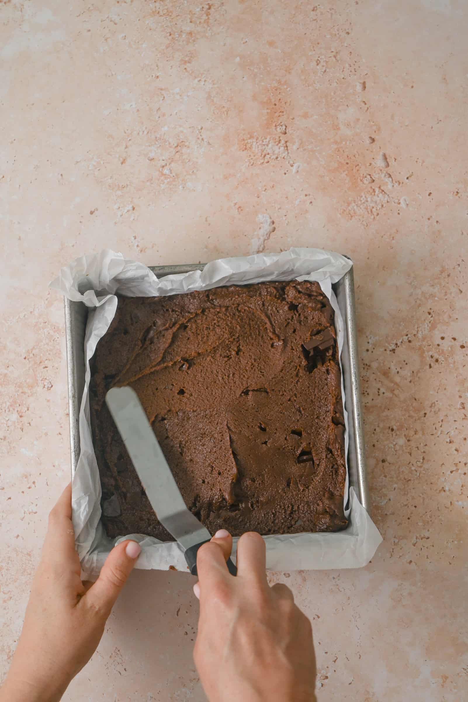 A person spreads chocolate batter evenly in a square baking pan lined with parchment paper, using a metal spatula, on a light brown countertop.