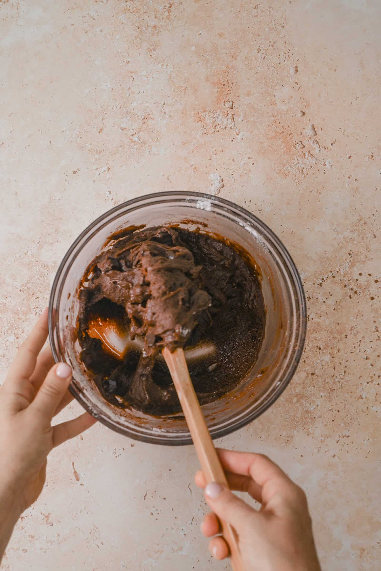 A person mixing chocolate batter in a glass bowl with a spatula on a light, textured surface.