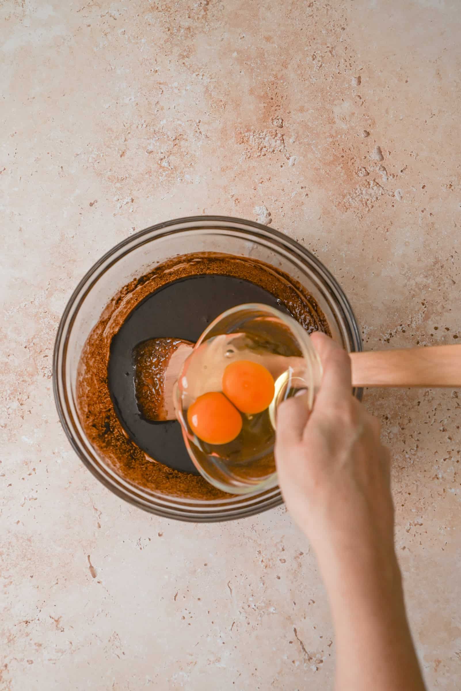 A hand holds a small bowl with two raw eggs above a glass mixing bowl containing a brown batter and a spatula on a light countertop.