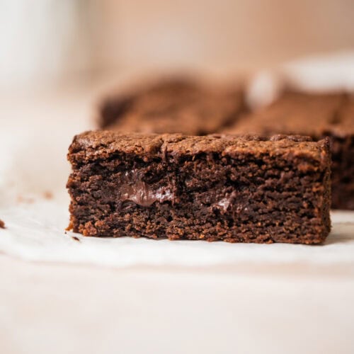 A close-up of a thick, fudgy chocolate cookie bar with a gooey chocolate center, sitting on white parchment paper.