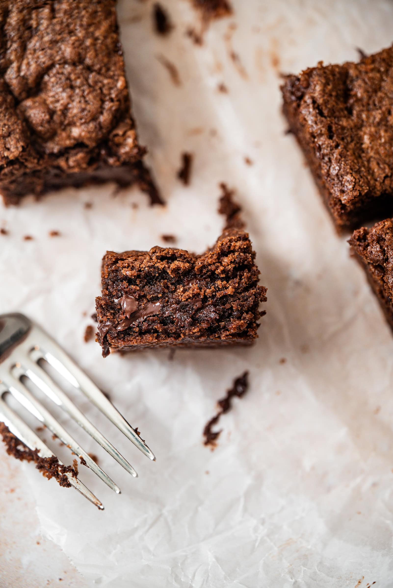 A close-up of a bitten brownie with a gooey chocolate center, surrounded by other brownies and a silver fork on crinkled parchment paper.