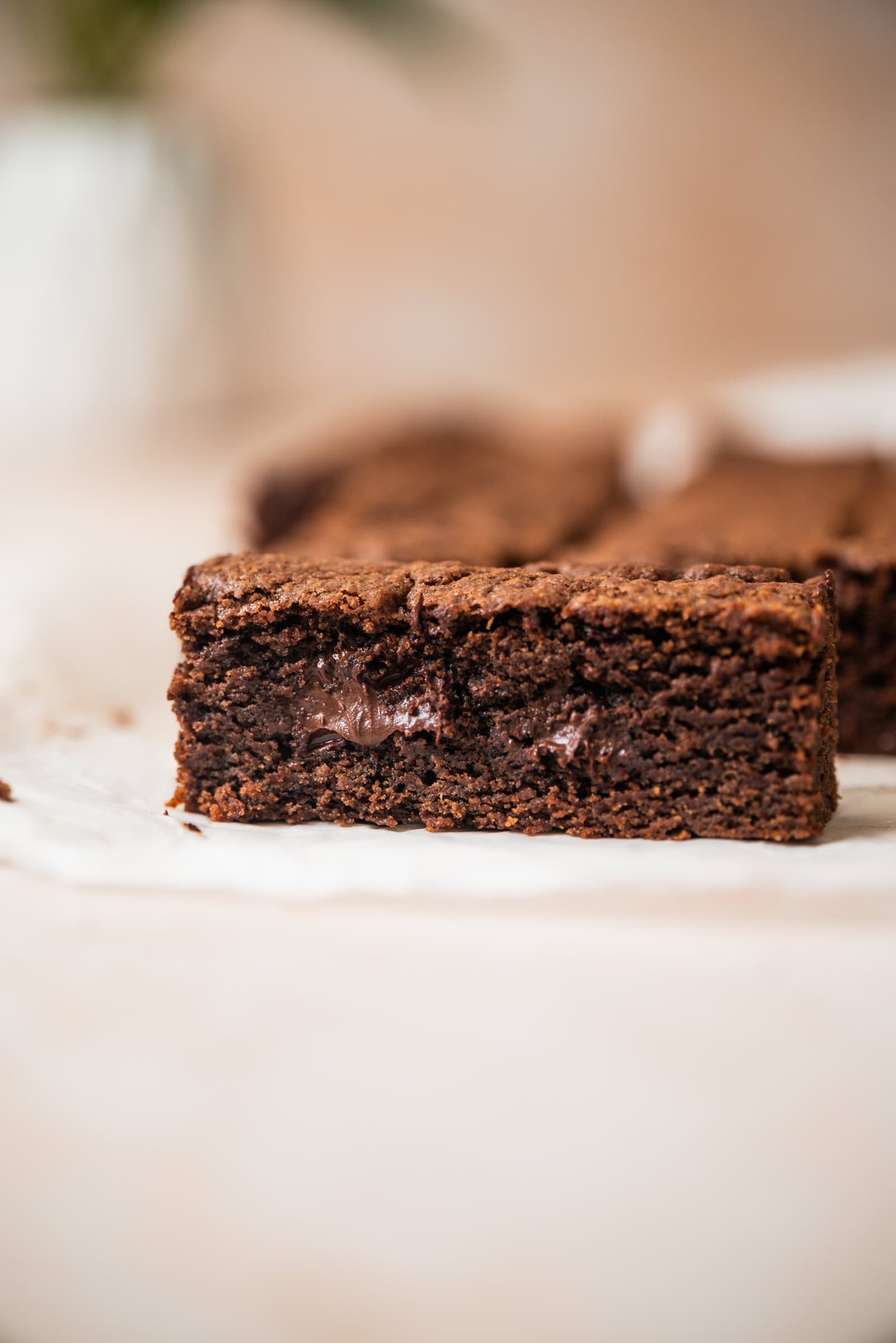 A close-up of a thick, fudgy chocolate cookie bar with a gooey chocolate center, sitting on white parchment paper.