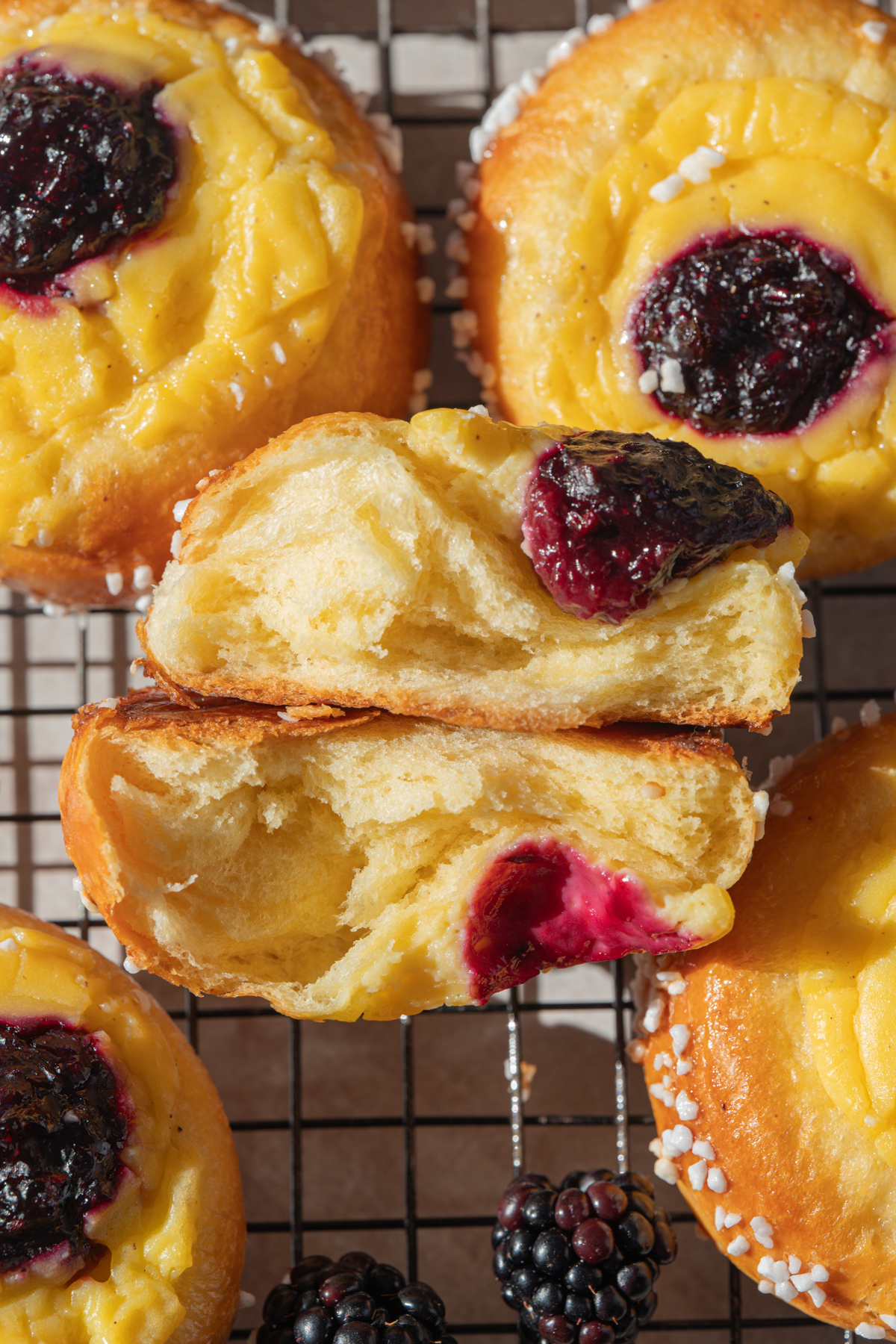 Close-up of blackberry and custard pastries on a cooling rack. One pastry is split open, showing its soft, airy inside. Fresh blackberries are arranged at the bottom of the image.