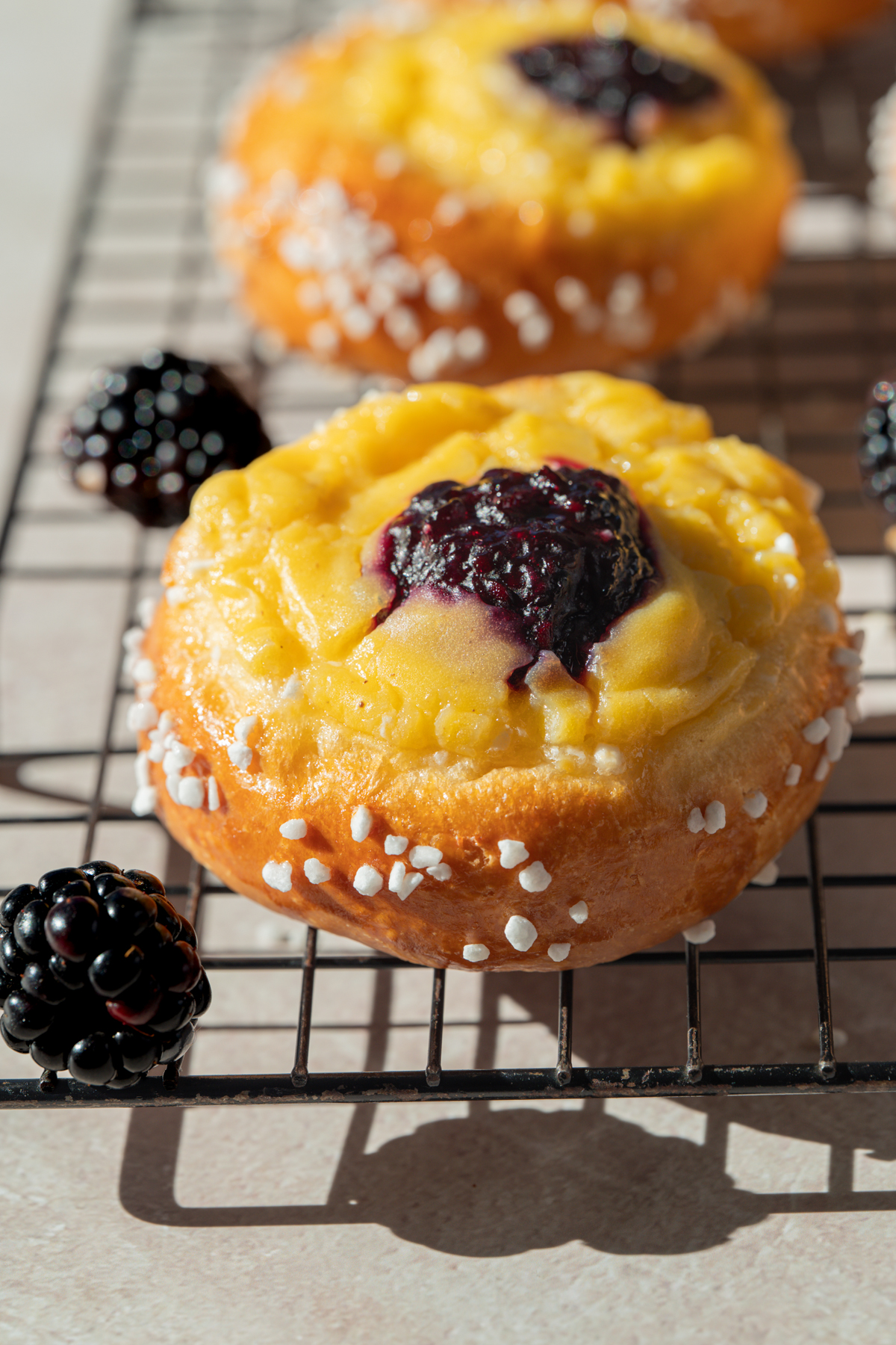 A close-up of a pastry topped with yellow custard and blackberry jam, sprinkled with coarse sugar, on a cooling rack with blackberries nearby.