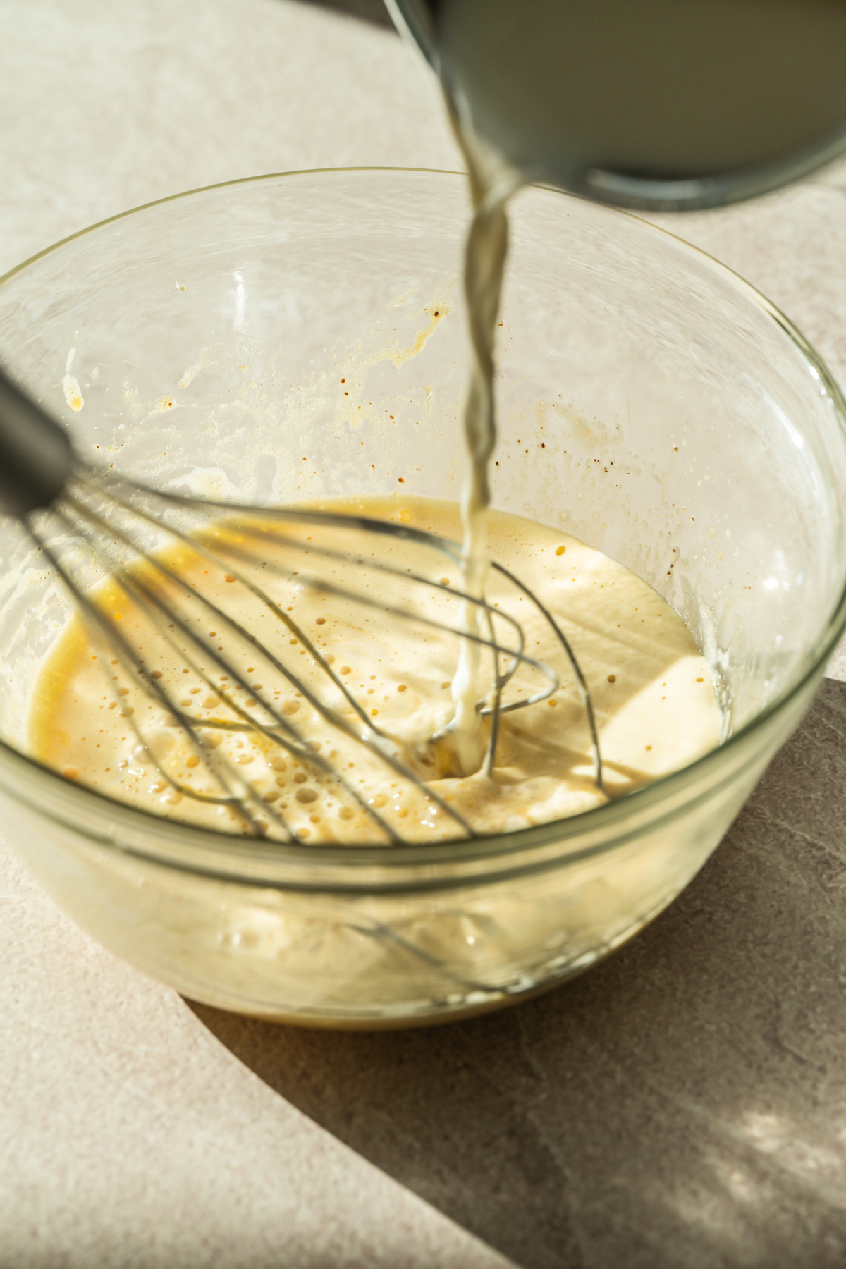 A close-up of milk being poured into a glass bowl containing a beaten egg mixture, with a metal whisk resting inside the bowl on a light countertop.
