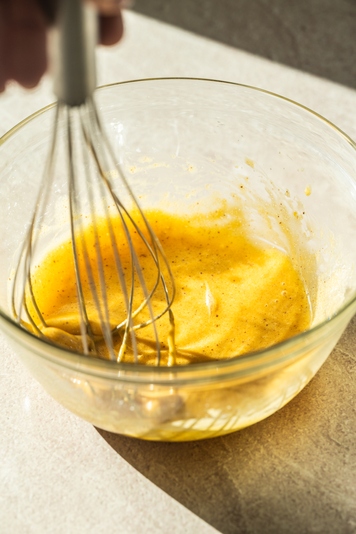 A hand whisking a yellow, frothy mixture in a clear glass bowl on a light-colored countertop, with sunlight streaming across the scene.