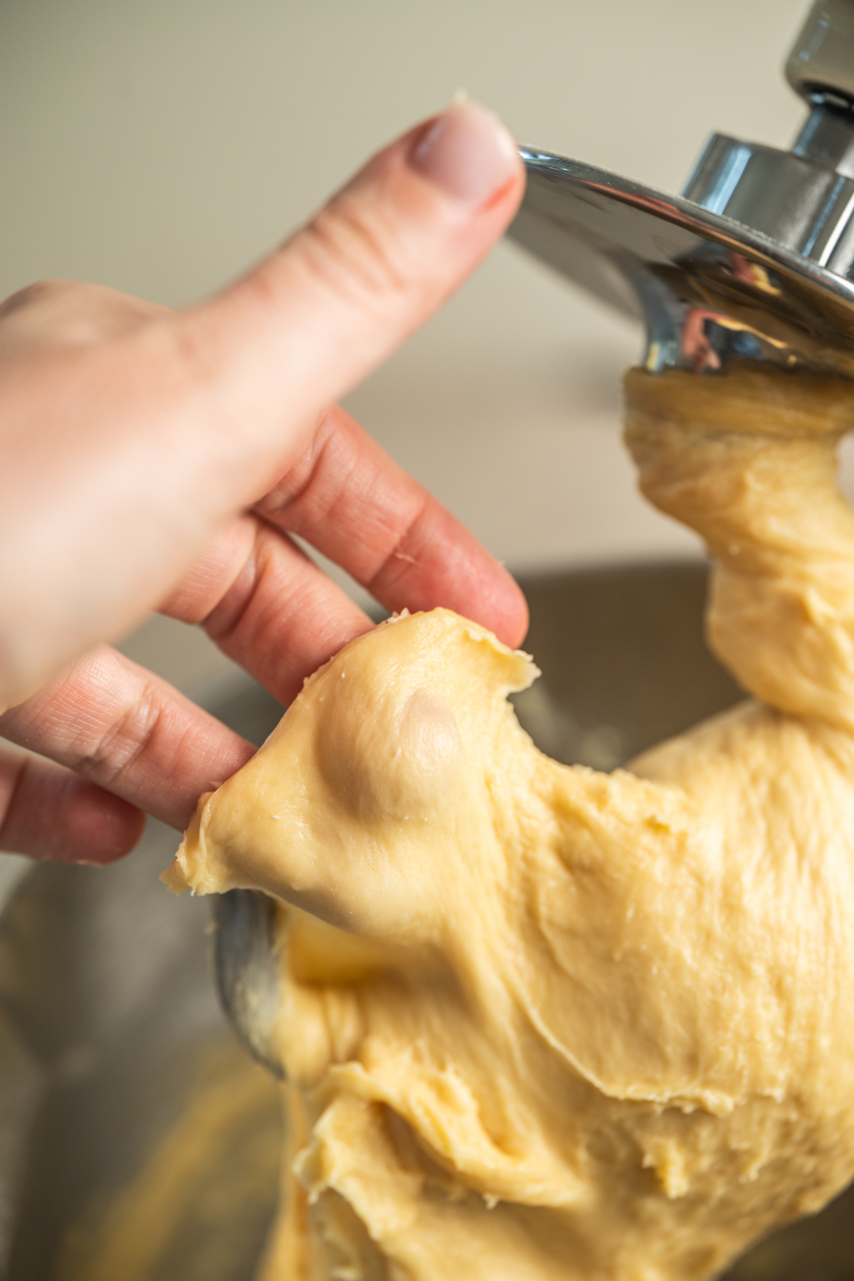 A hand stretches a piece of smooth, elastic dough from a stand mixer hook, showing its texture and readiness for baking.