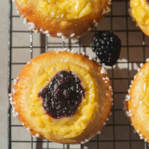 Brioche pastries topped with yellow custard and blackberry jam, placed on a cooling rack with fresh blackberries scattered around.