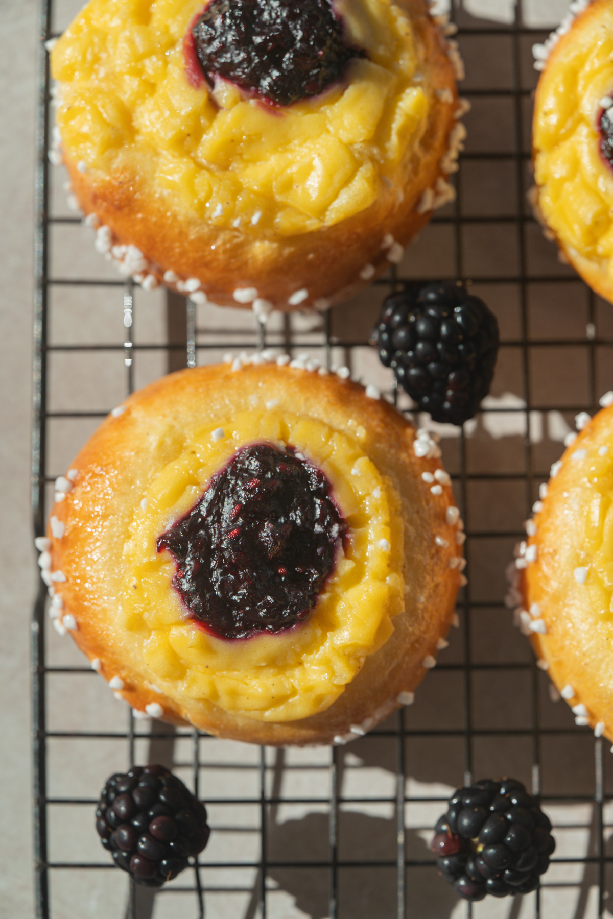 Brioche pastries topped with yellow custard and blackberry jam, placed on a cooling rack with fresh blackberries scattered around.