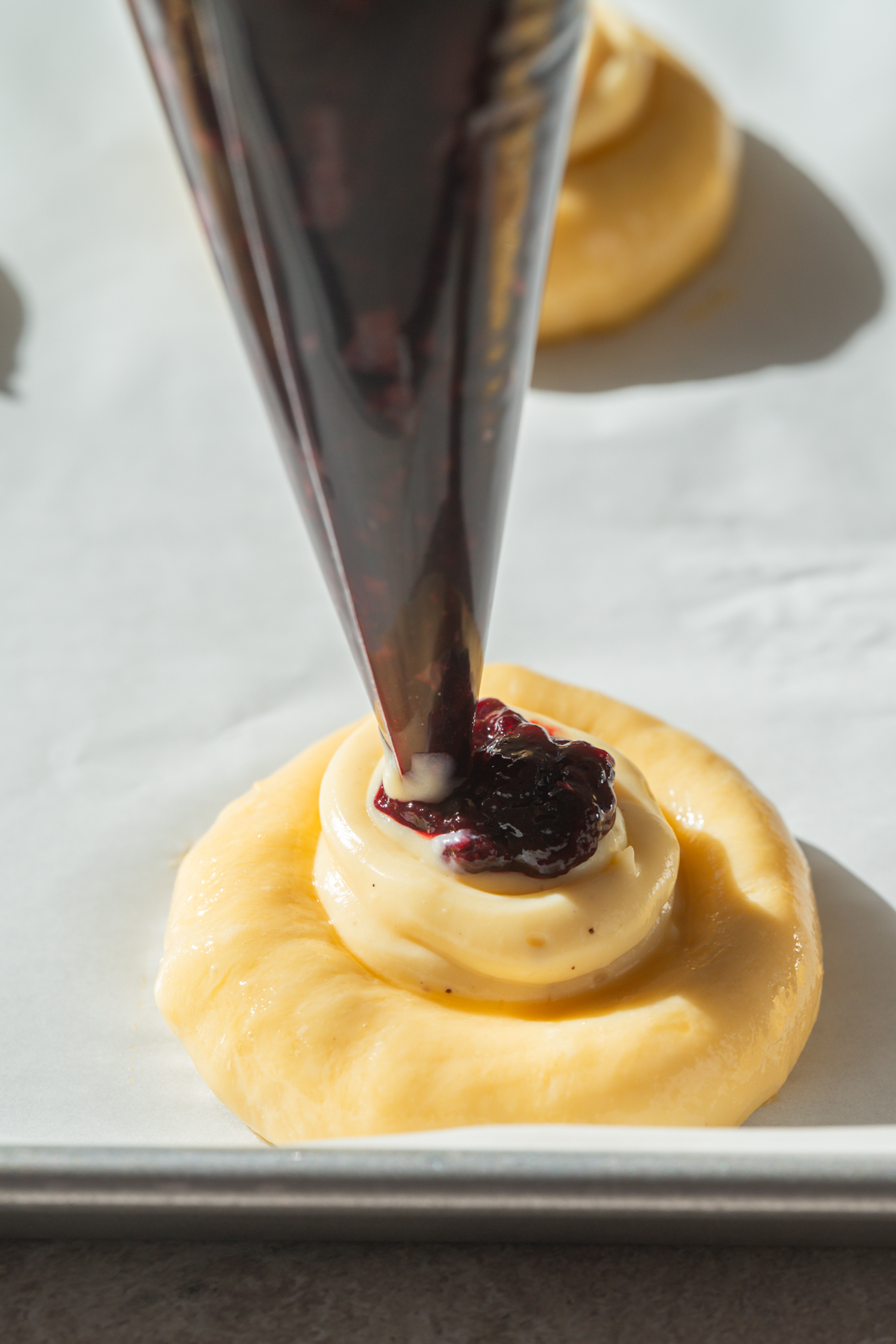 A close-up of a pastry being filled with cream, as a piping bag adds a dollop of dark berry jam onto the cream, all on a baking sheet lined with parchment paper.