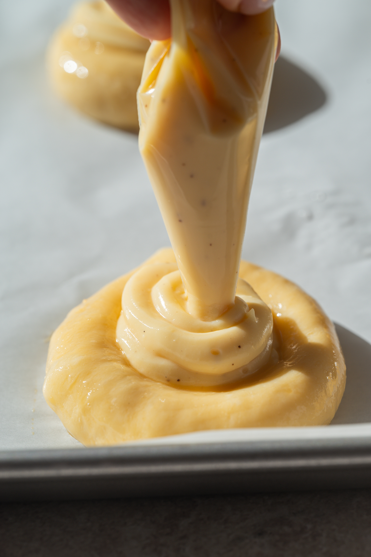 A hand pipes creamy filling onto a round pastry dough, both sitting on a parchment-lined baking sheet in bright natural light.