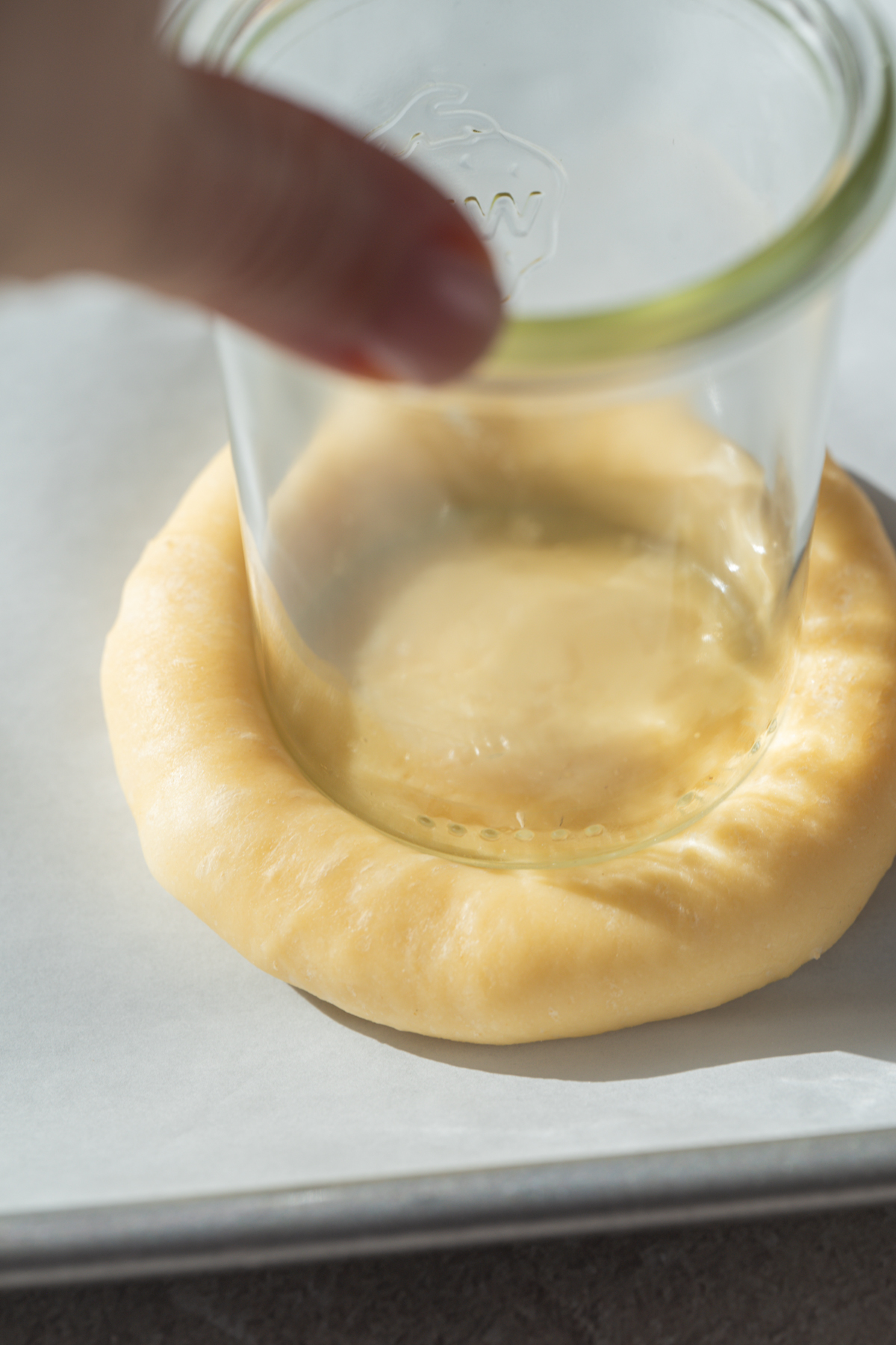 A hand presses a glass jar into the center of a round piece of dough on a baking sheet, creating an indentation while leaving a raised edge.