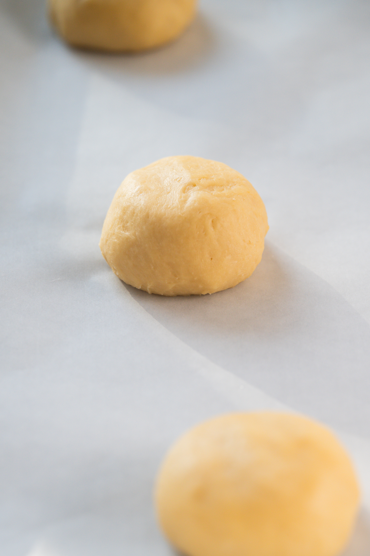 A ball of raw cookie dough sits on a sheet of parchment paper, with another dough ball blurred in the foreground.