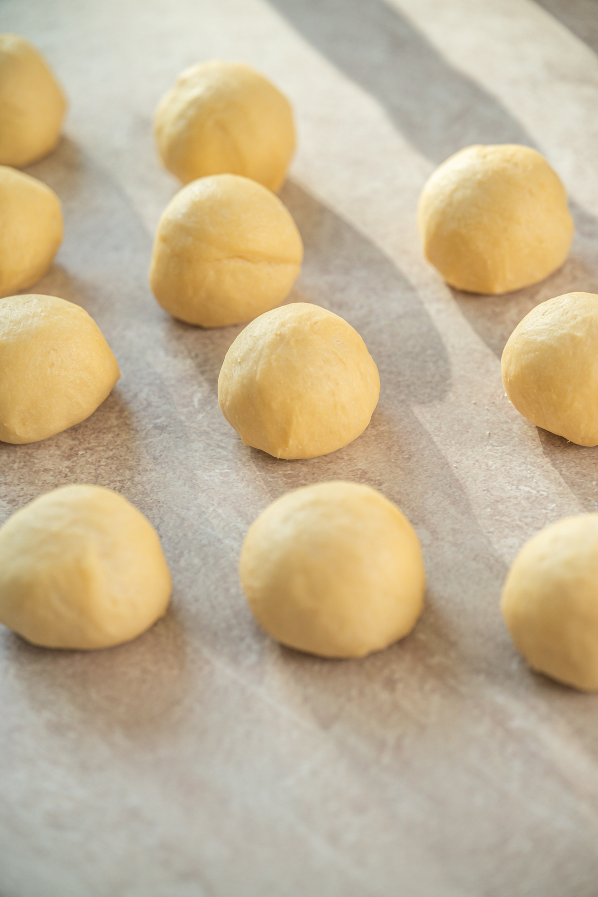 Several smooth, round balls of dough are evenly spaced on a lightly floured surface, ready for baking. The sunlight creates soft shadows across the dough balls.