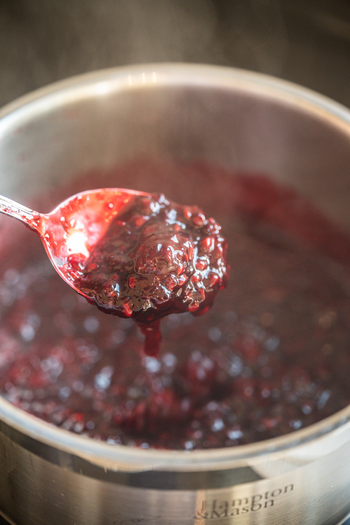 A close-up of a metal spoon holding a thick, dark red berry jam or sauce over a stainless steel pot filled with the same mixture. Steam rises from the pot, indicating it is hot.