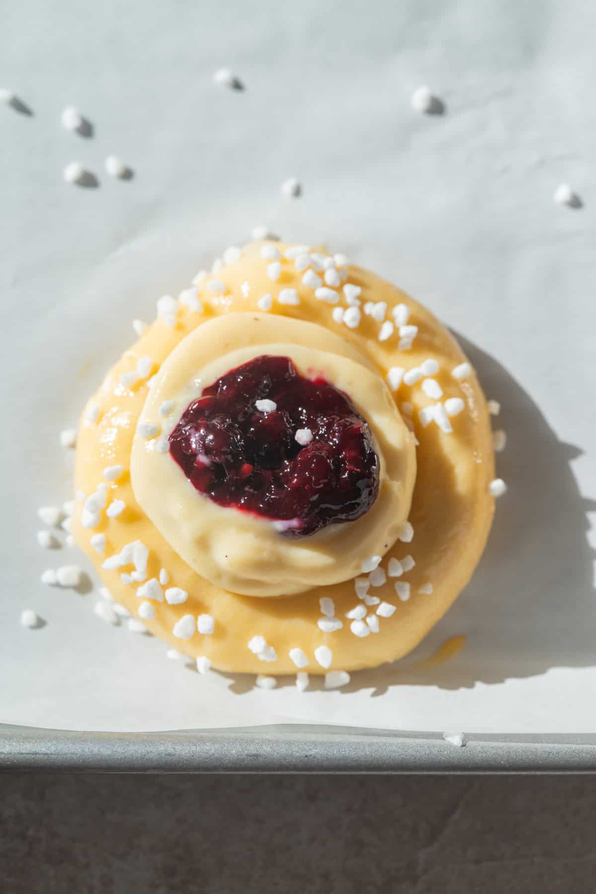 A close-up of an unbaked pastry topped with vanilla custard, a dollop of berry jam in the center, and sprinkled with pearl sugar, placed on parchment paper.